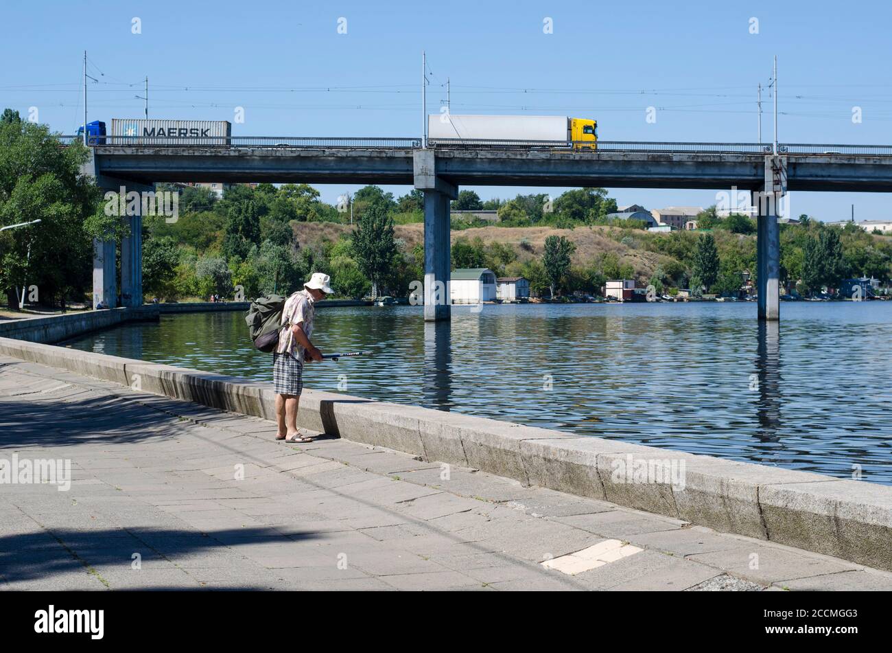 Nikolaev, Ucraina - 17 agosto 2020: L'uomo maturo cattura il pesce sull'argine della città. I trattori per camion attraversano il ponte sul fiume Ingul nel bac Foto Stock