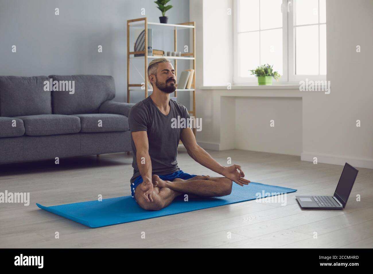Lezione di yoga online. Giovane ragazzo tranquillo che pratica la meditazione mattutina di fronte al computer portatile a casa. Concetto di equilibrio di vita Foto Stock