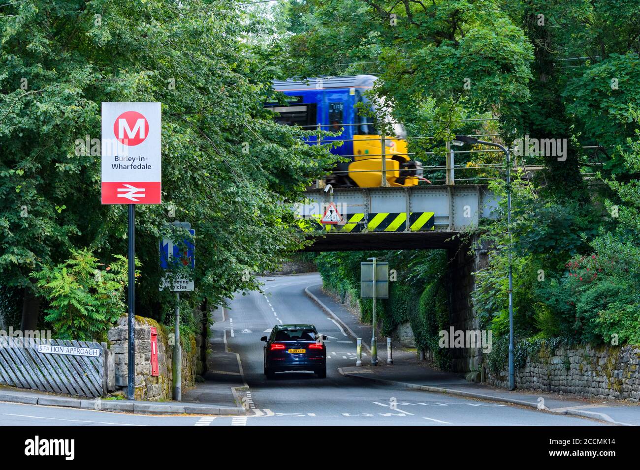 Treno di servizio passeggeri della Northern Rail, passando l'insegna della stazione sul ponte ferroviario basso e auto sulla strada rurale del villaggio - Burley, Yorkshire, Inghilterra, Regno Unito Foto Stock