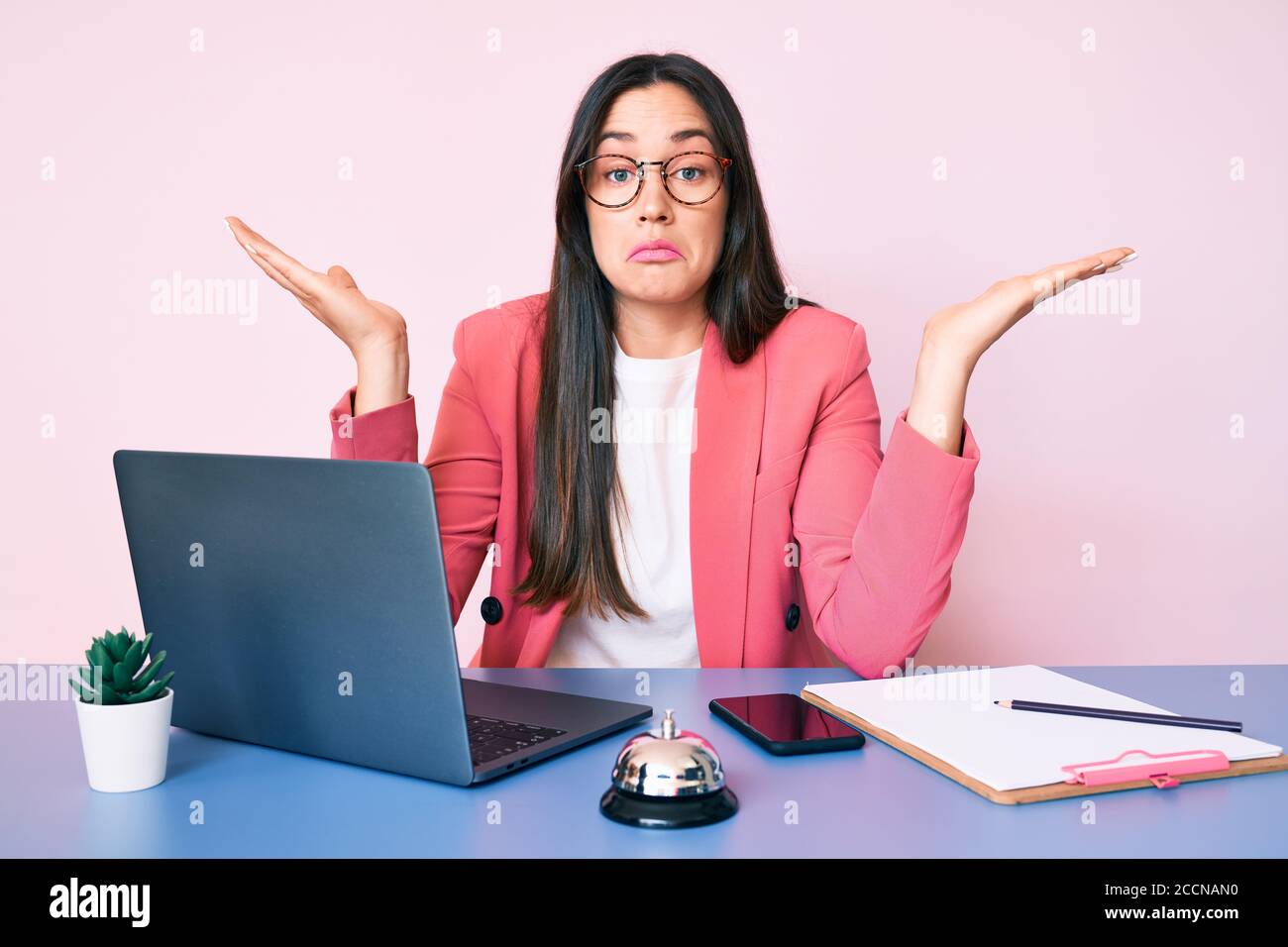 Giovane donna caucasica seduta alla scrivania del receptionist che lavora usando il laptop indeceless e l'espressione confusa con le braccia e le mani sollevate. Concetto di dubbio Foto Stock
