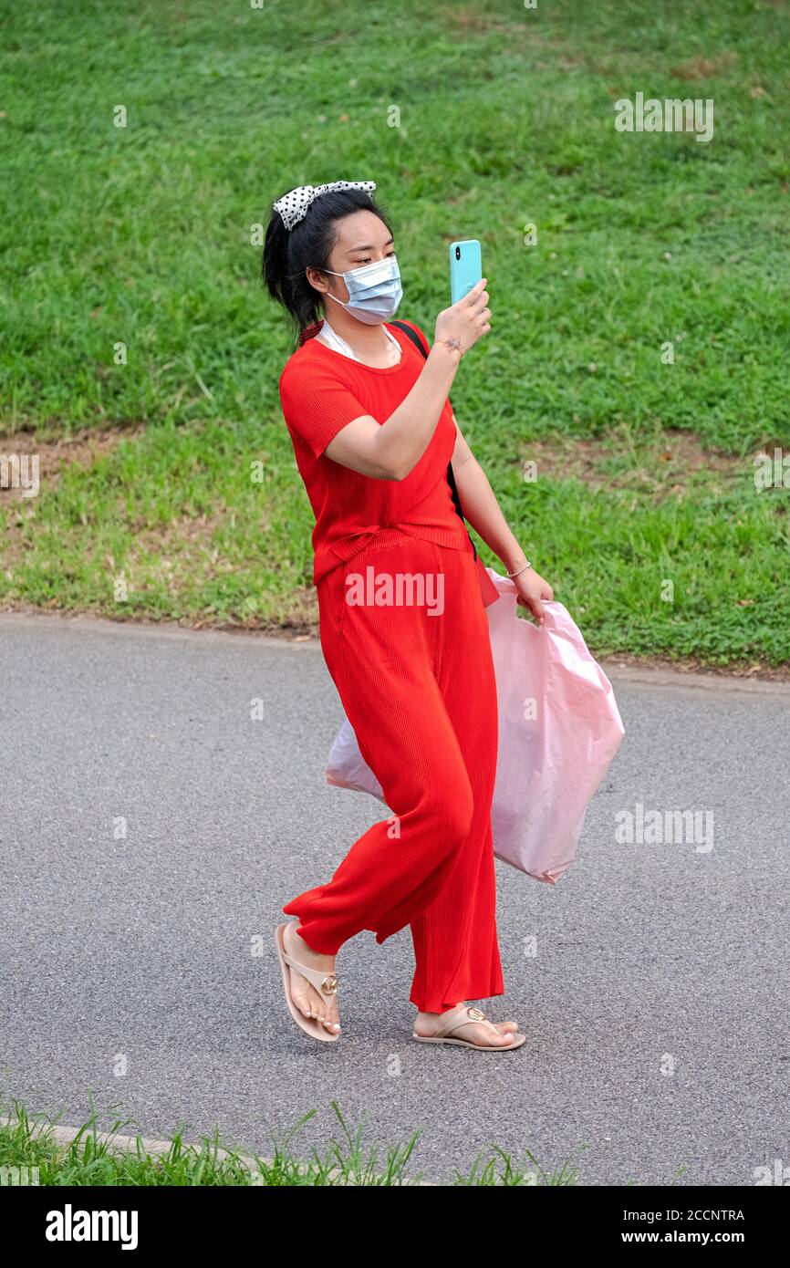 Una donna che indossa una tuta da pantaloni rossi e un arco di capelli a pois di polka legge il suo telefono cellulare mentre cammina in un parco a Queens, New York City. Foto Stock