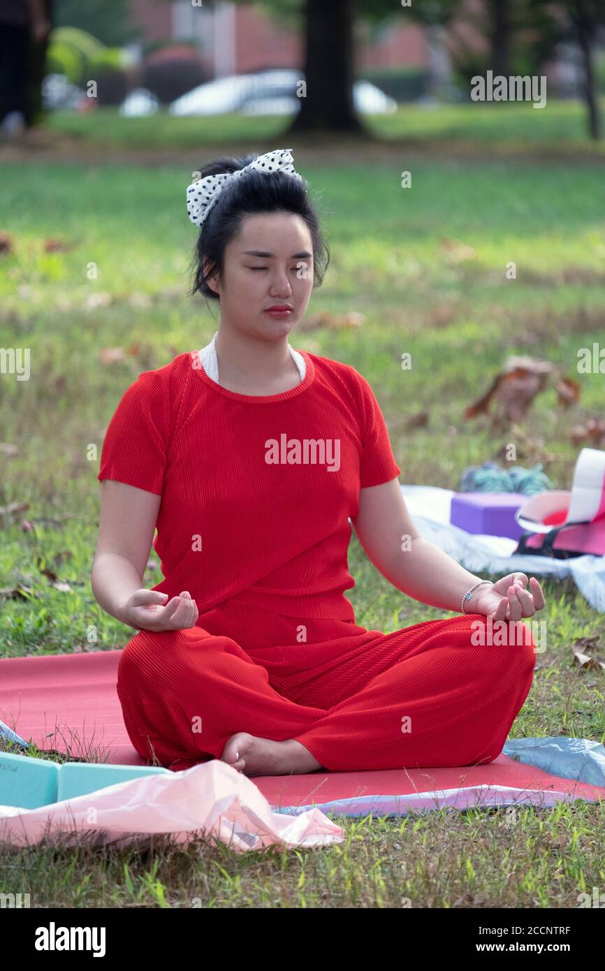 Una donna che indossa una tuta con pantaloni rossi e un arco a pois di pois si siede nella posizione del loto ad una classe di yoga in un parco a Queens, New York Foto Stock
