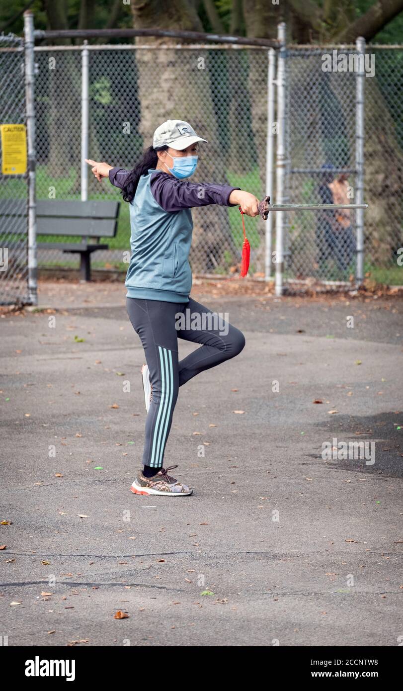 Una donna asiatica americana che indossa una maschera chirurgica che lavora con una spada di Shaolin durante una lezione di Tai Chi in un parco a Flushing, Queens, New York Foto Stock