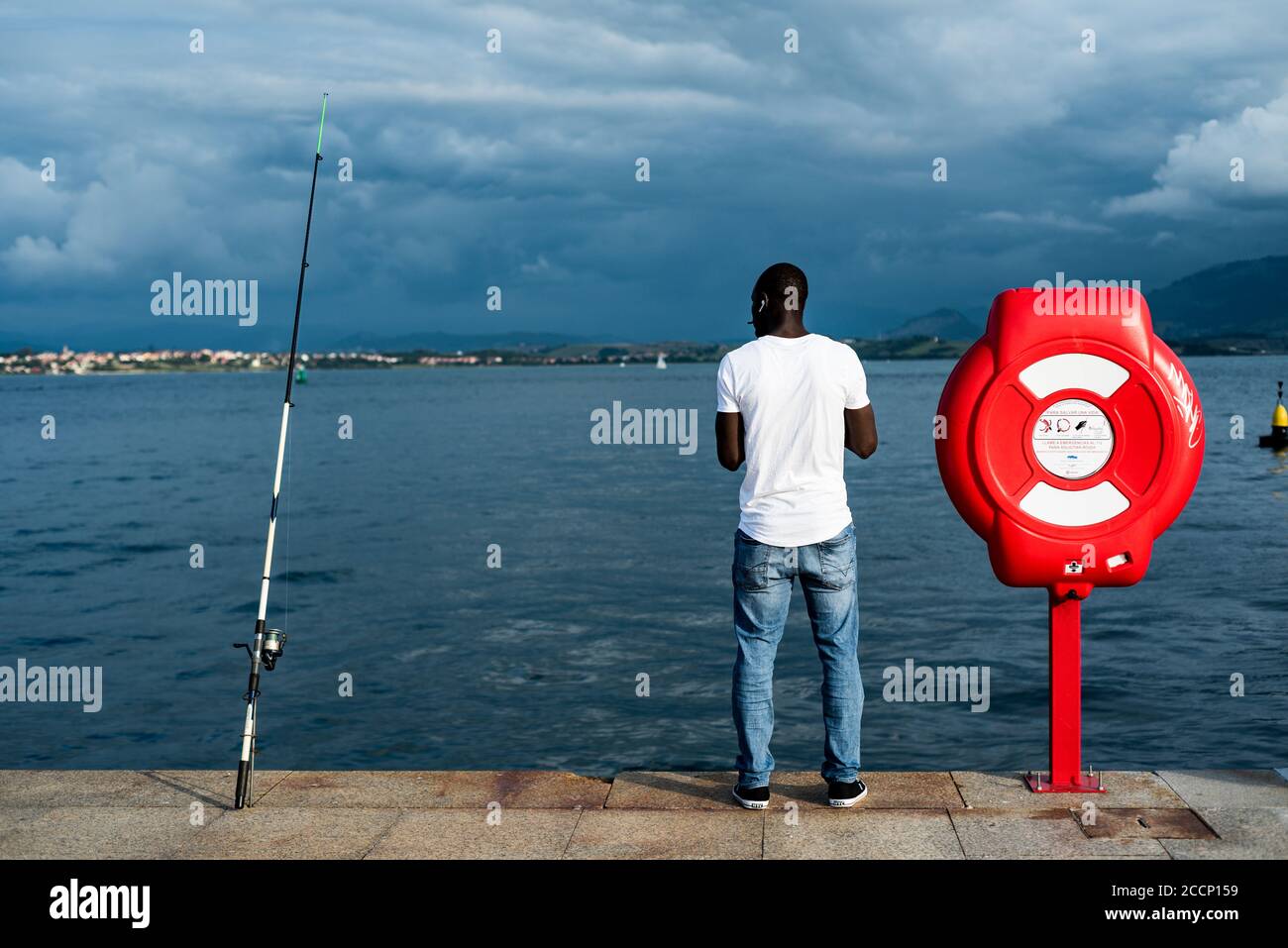 Vista posteriore di un giovane uomo nero che indossa una T-shirt bianca. Uomo nero che ascolta la musica mentre pesca in Paseo Maritimo, Santander, Spagna Foto Stock