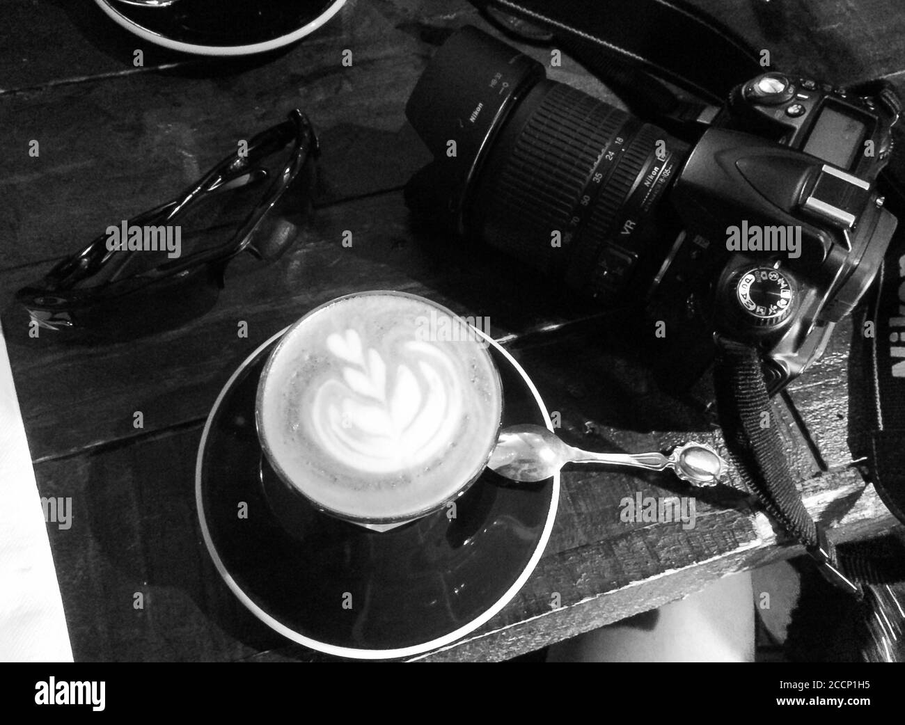 Foto in bianco e nero di una tazza di cappuccino e fotocamera DSLR in un bar. Foto Stock