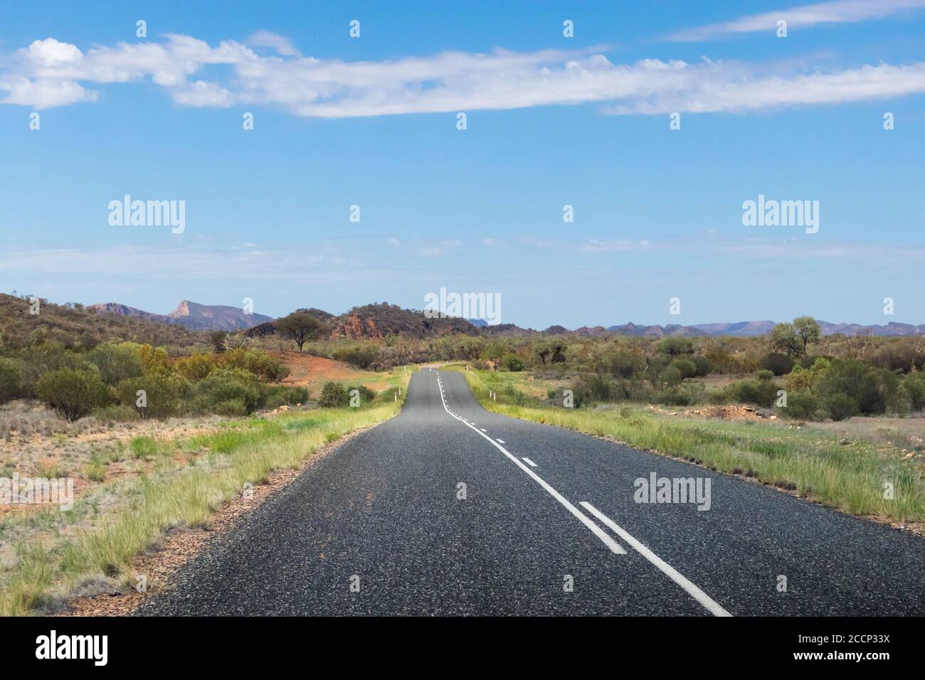 Guida attraverso le MacDonnell Ranges. Montagne sullo sfondo. Strada vuota, senza auto, senza segnaletica. Vegetazione verde e cespuglio. Australia Foto Stock