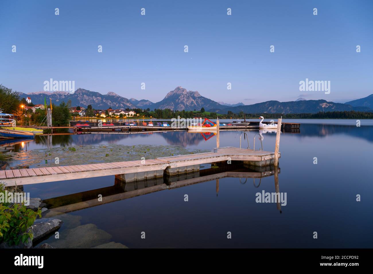 Pontile in legno a Hopfensee, sullo sfondo le Alpi e il villaggio di Hopfen am See, Baviera, Germania Foto Stock