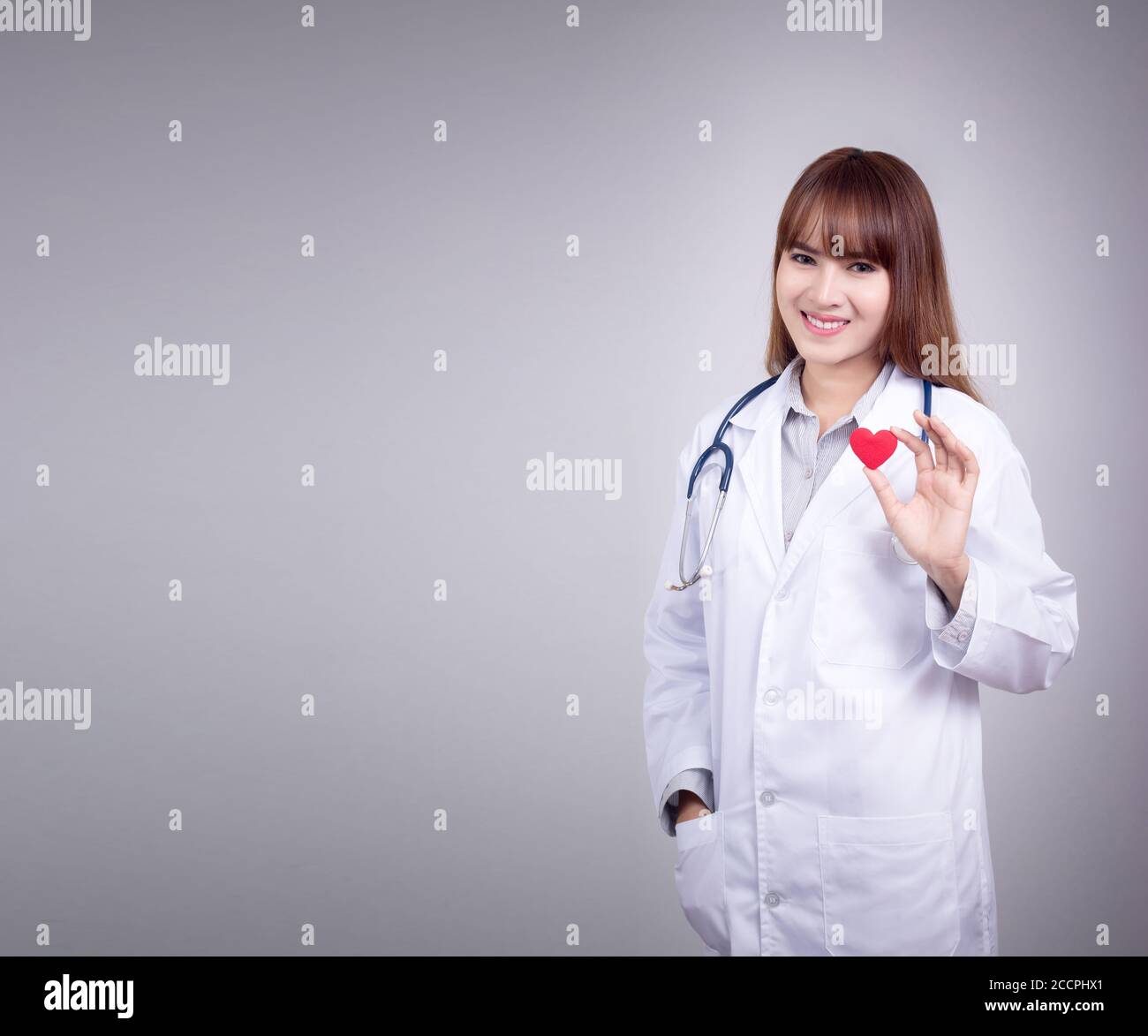 Giovane medico asiatico stare tenendo un cuore rosso in mano con faccia sorridente. Vesti con uniforme di abito medico con stetoscopio in un ospedale. Bellissima Foto Stock