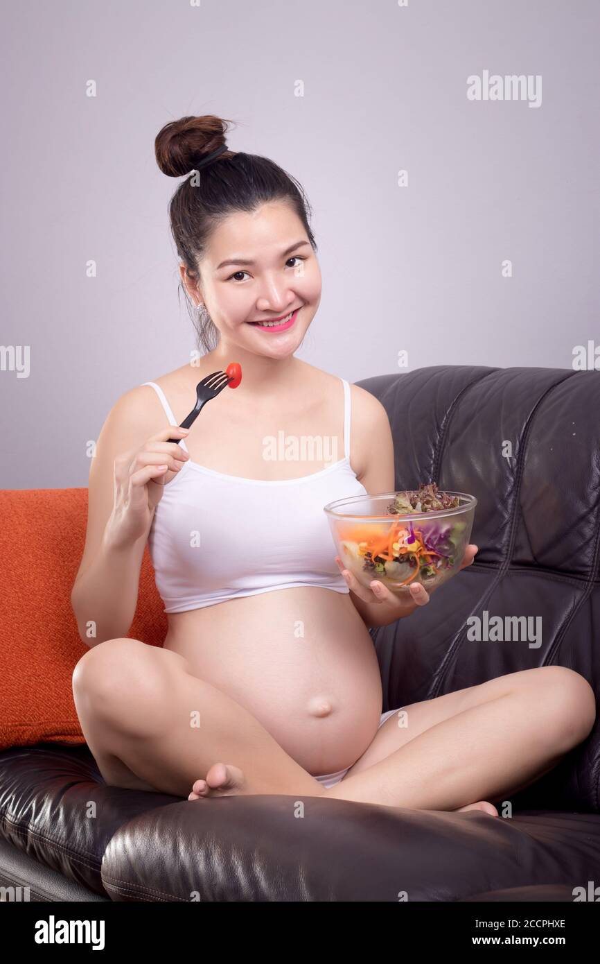 Concetto sano di nutrizione incinta. Felice giovane bella donna in gravidanza asiatica mangiare insalata da ciotola insalata sul divano con faccia sorridente. Assistenza sanitaria con re Foto Stock