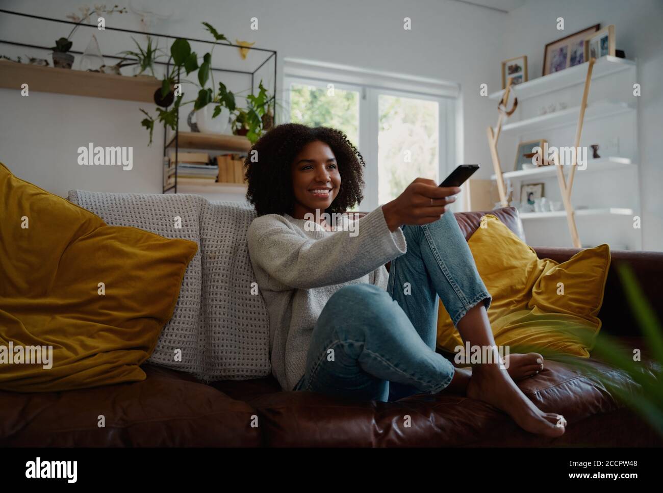 Giovane donna seduta in interni guardando la tv e tenendo il telecomando sul divano Foto Stock
