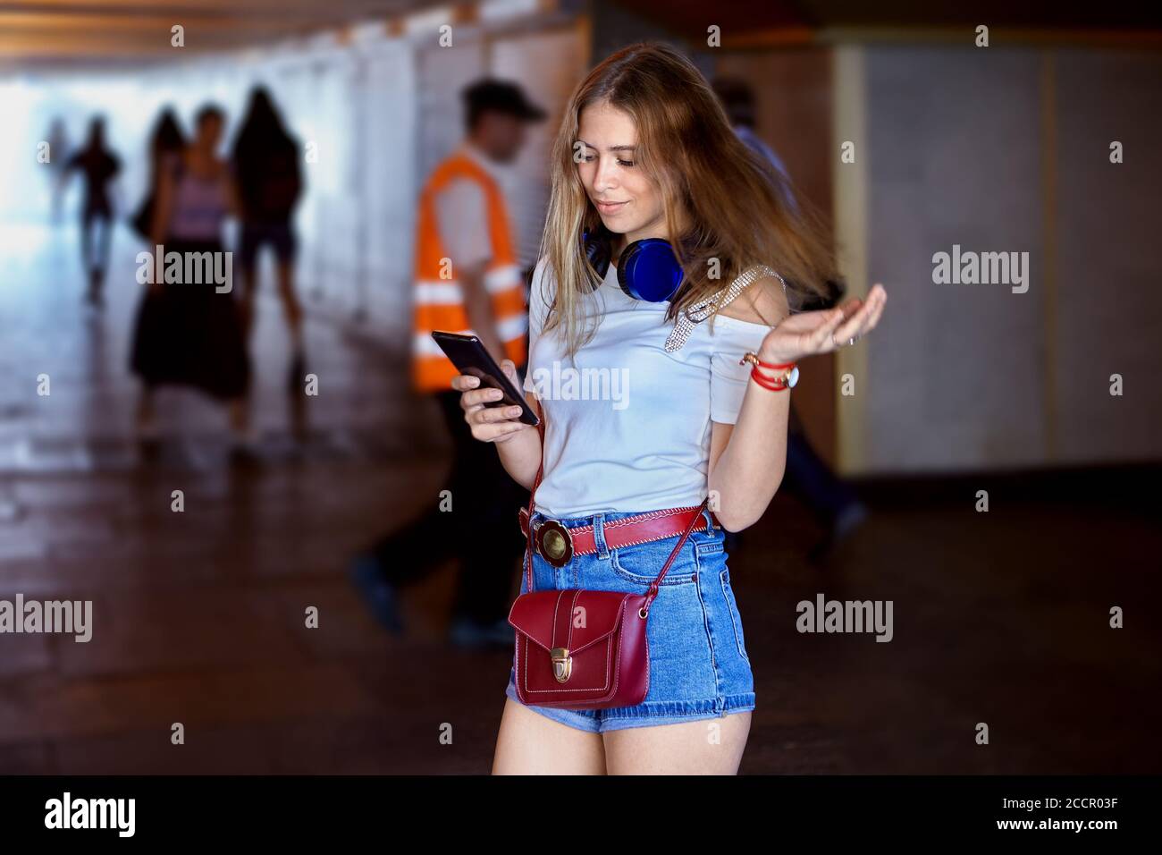 Attraente giovane donna caucasica di circa 25 anni con lungo capelli biondi e auricolari e vestiti in un panno alla moda è in piedi in sottopassaggio con Foto Stock