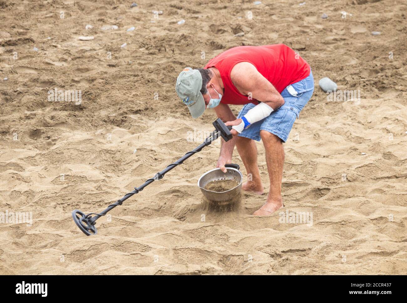 Uomo che indossa la copertura del viso, maschera facciale che usa il metal detector sulla spiaggia durante la pandemia di Covid/Coronavirus. Foto Stock