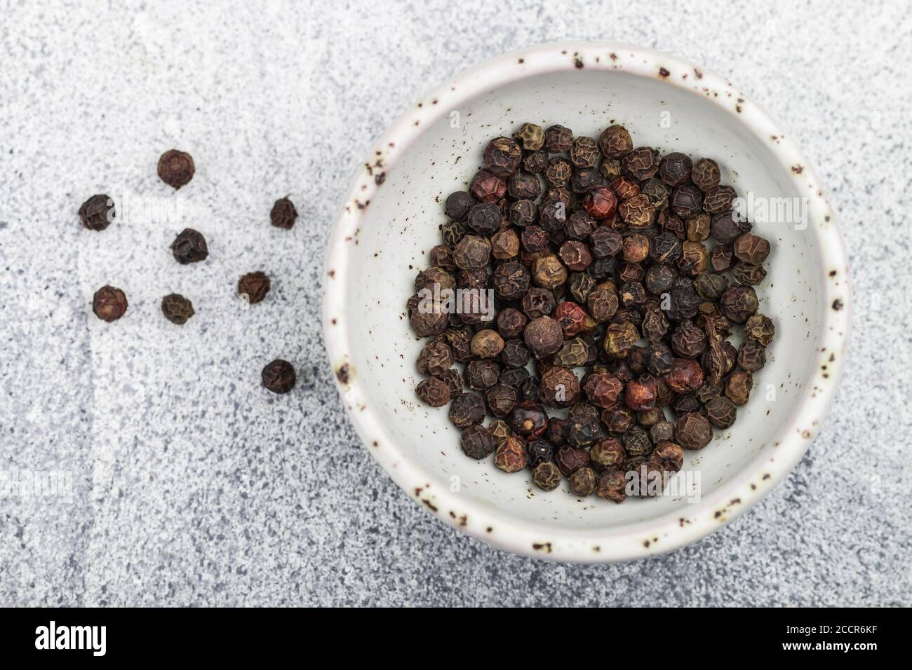 Grani di pepe secchi organici neri in una ciotola bianca su un calcestruzzo leggero o un primo piano di fondo di pietra. Spezie e condimenti. Messa a fuoco selettiva, vista dall'alto e. Foto Stock