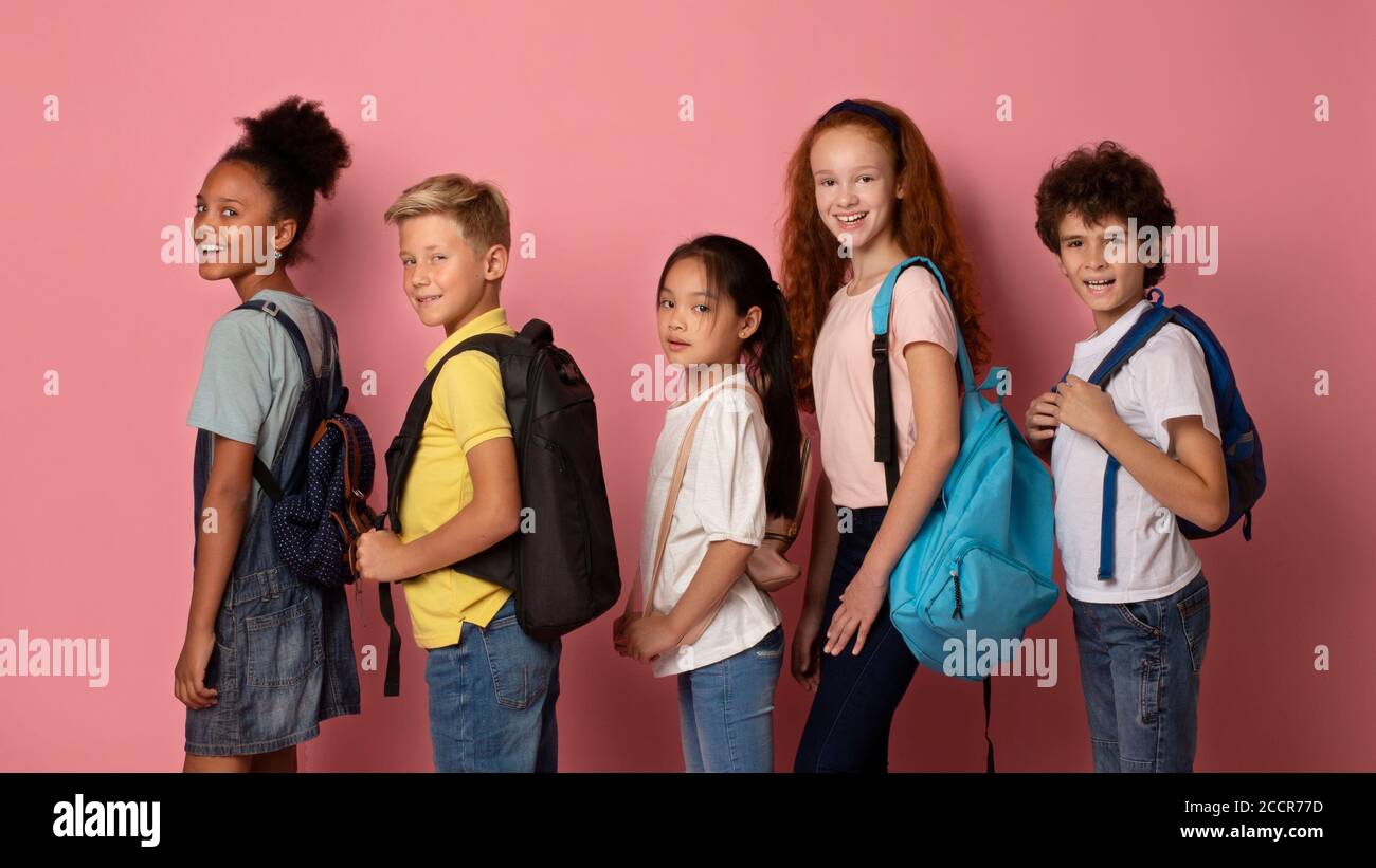 Bambini gioiosi con zaini che vanno a scuola uno dopo l'altro su sfondo rosa Foto Stock