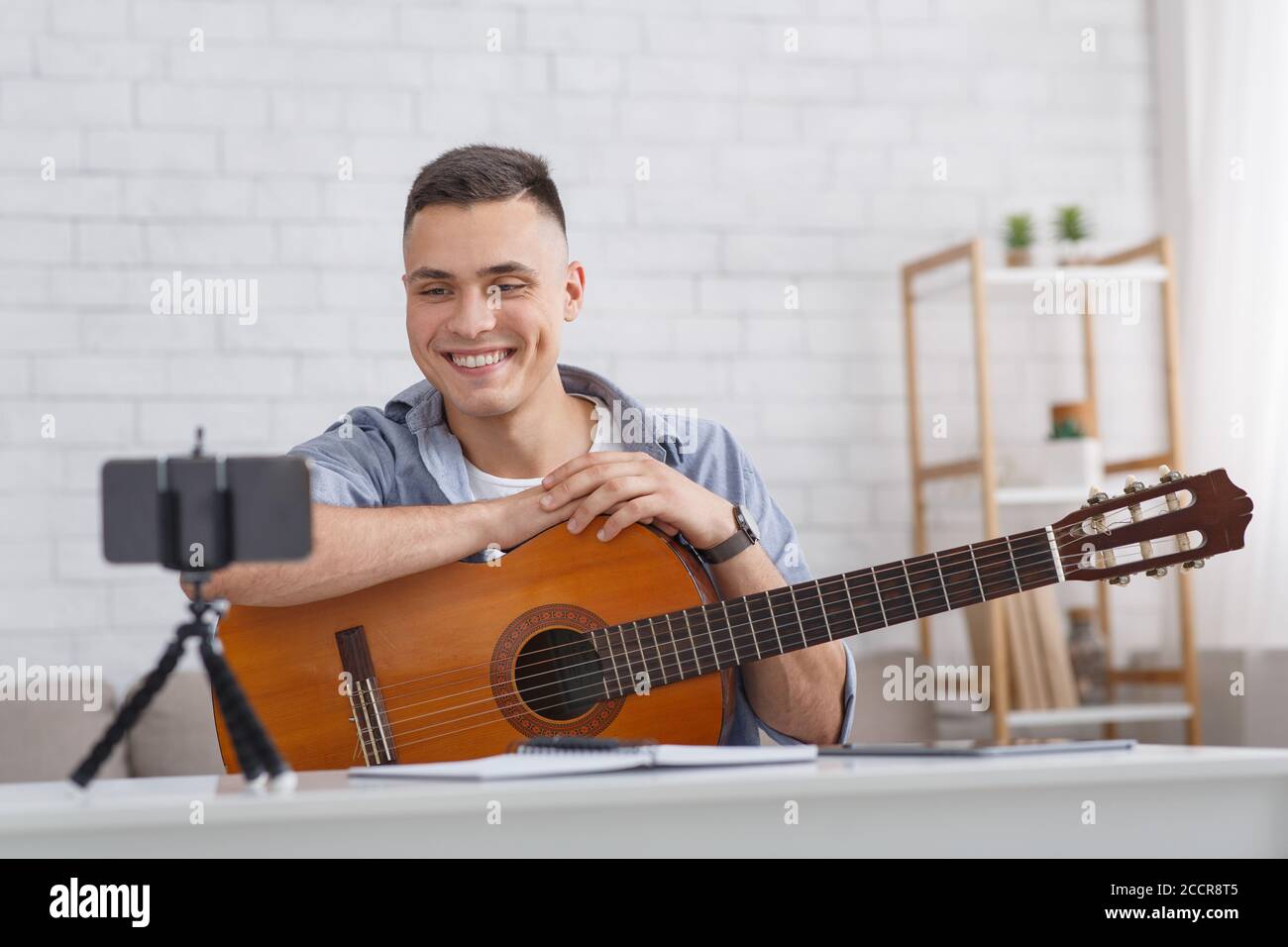 Lezione di musica online. Un ragazzo sorridente con la chitarra guarda alla fotocamera dello smartphone Foto Stock