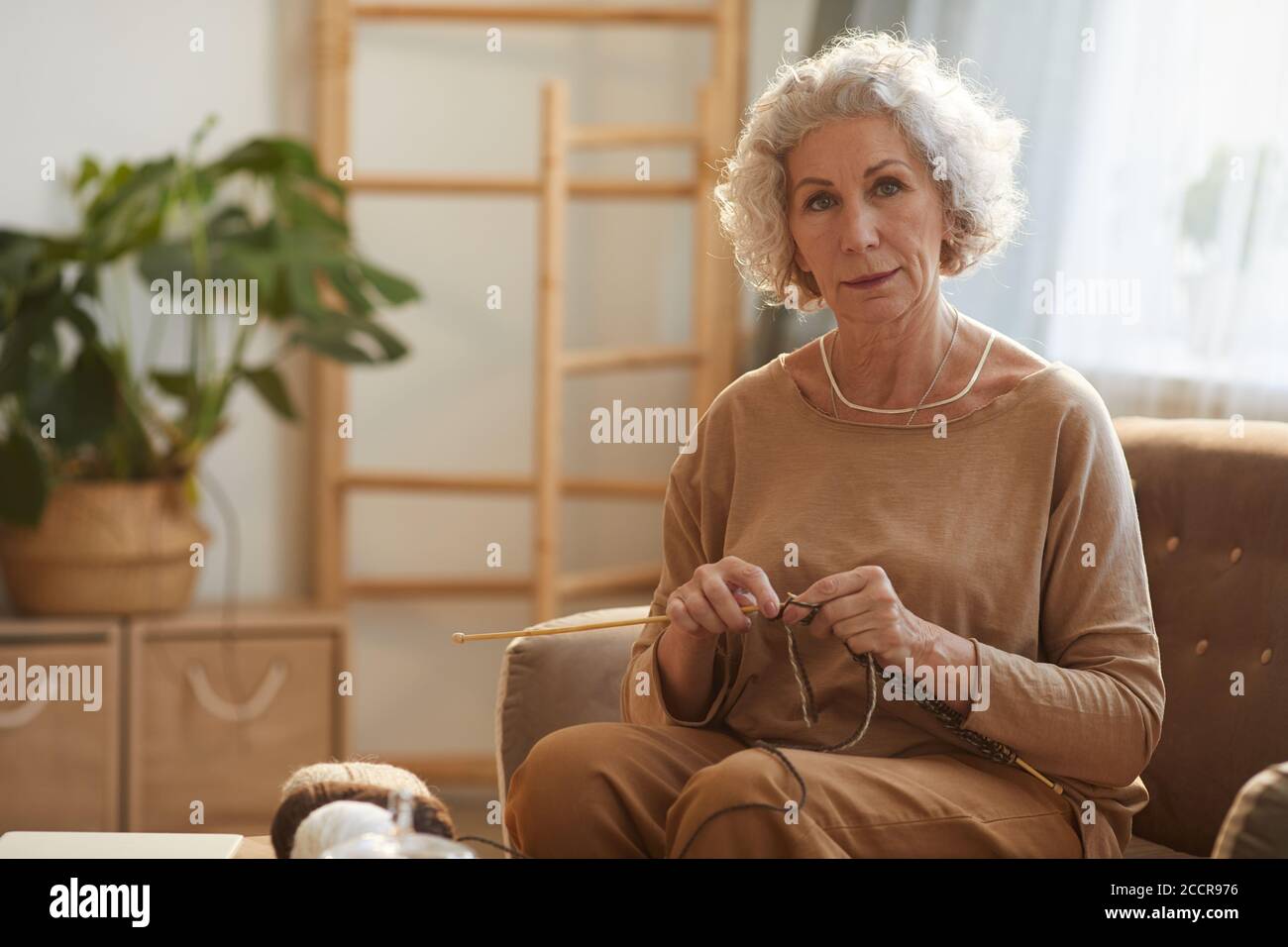 Un ritratto dai toni caldi di un'elegante donna anziana che si accovona e guarda la fotocamera mentre si siede sul divano in una casa accogliente illuminata dalla luce del sole, spazio per la copia Foto Stock