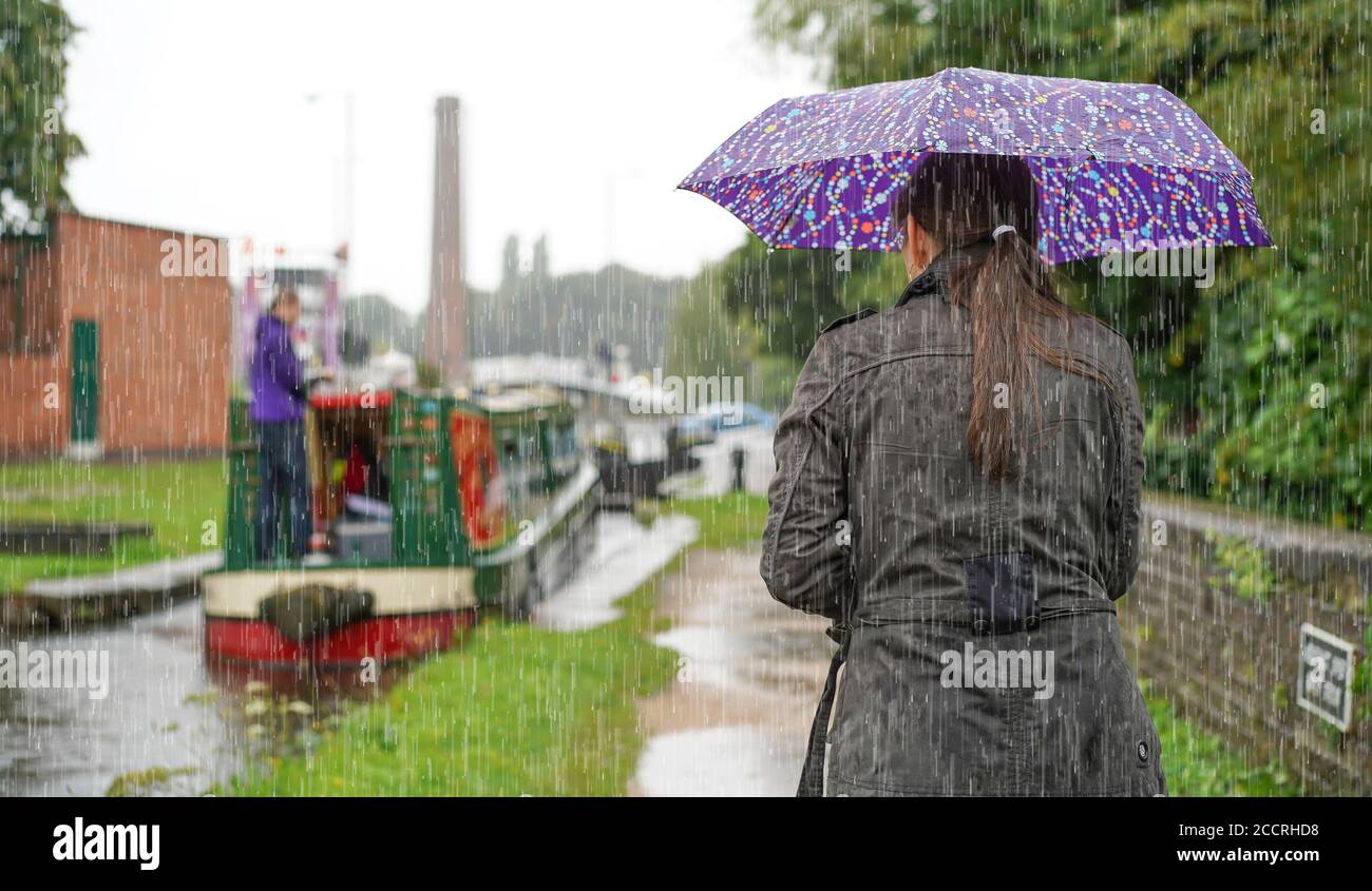 Donna da dietro (corpo superiore) che ripara sotto l'ombrello in piedi sotto la pioggia torrenziale, guardando narrowboat sul canale britannico che si avvicina al blocco canale. Foto Stock