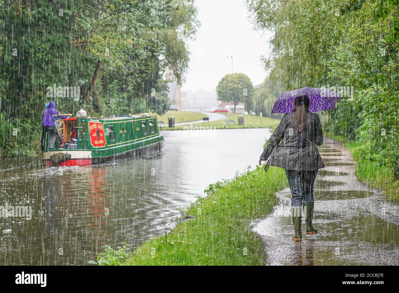 Vista posteriore di donna in salvi al riparo sotto l'ombrello camminando sull'alzaia lungo il narrowboat in movimento sul canale britannico in forte pioggia estiva britannica. Foto Stock