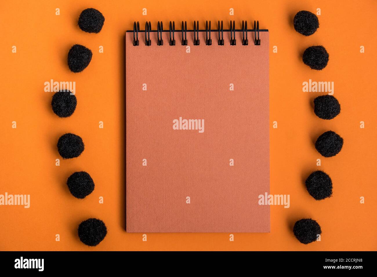 Blocco note arancione in una cornice di pom-pom nero soffice su uno sfondo arancione. Concetto di celebrazione di Halloween. Vista dall'alto, disposizione piatta. Spazio di copia. Bock up. Foto Stock