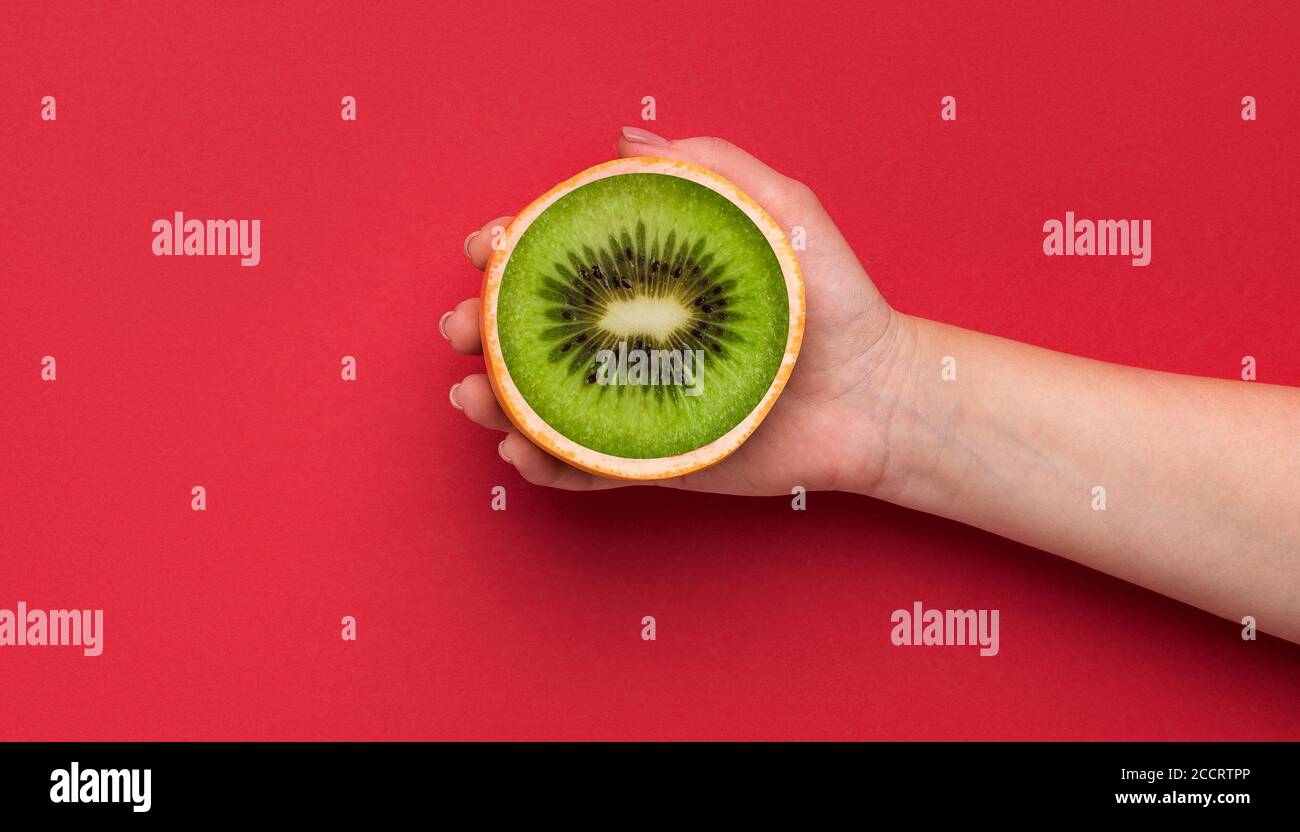 Mano femminile che tiene frutta arancione geneticamente modificata con centro kiwi su sfondo rosso, vista dall'alto. Collage Foto Stock