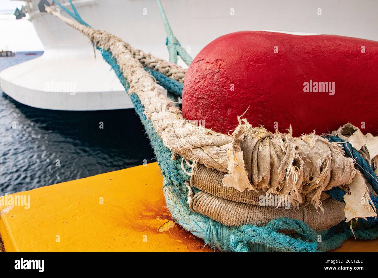 Corda nautica spessa e stagionato avvolta intorno al paletto rosso, che ormeggia una grande imbarcazione marina fino ad un molo/molo giallo brillante Foto Stock