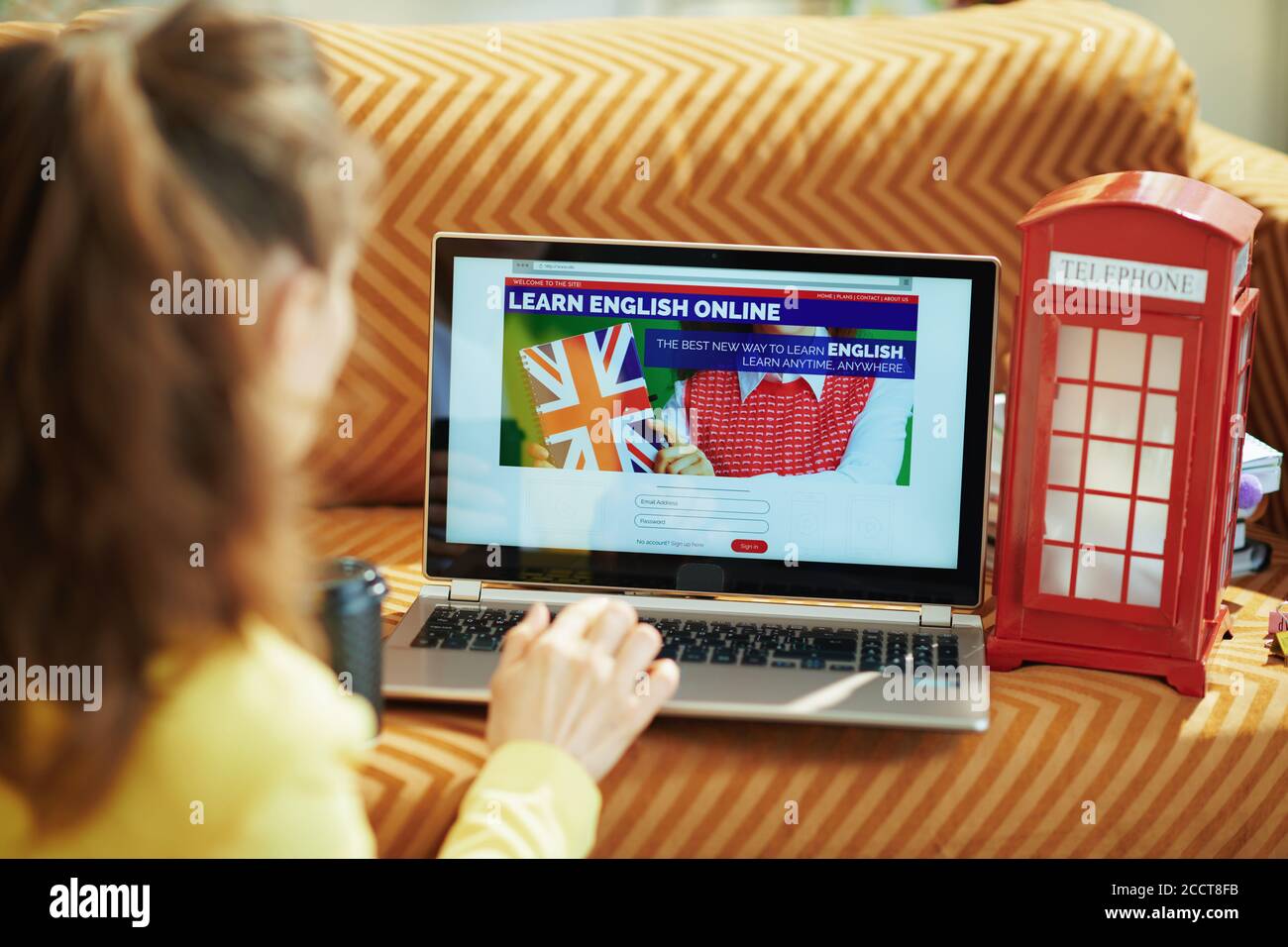 Visto da dietro la donna in casa in una giornata di sole imparando la lingua inglese online su un computer portatile. Foto Stock