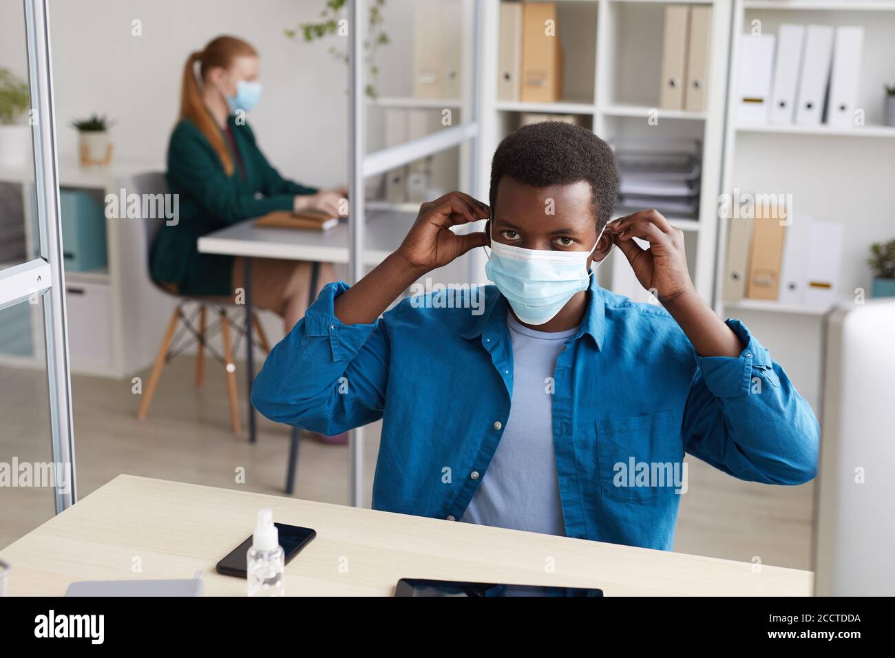 Ritratto ad alto angolo di un giovane afroamericano che si mette in faccia mascherare e guardare la fotocamera mentre si lavora alla scrivania ufficio postale pandemico Foto Stock