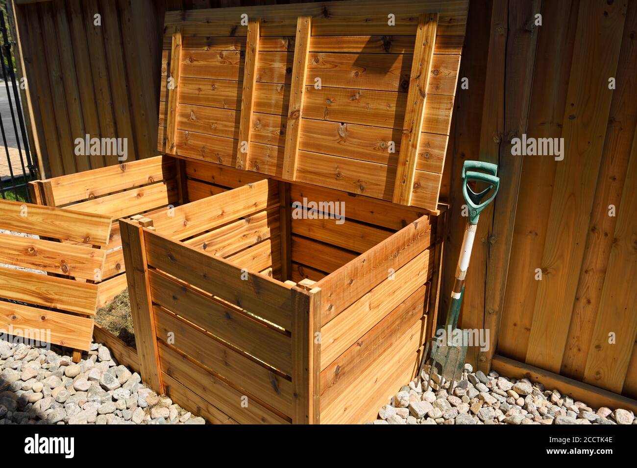 Due bidoni compostaggio organico in legno di cedro con aperto coperchio e rimosso parte anteriore per rifiuti di iarda accanto alla recinzione su rocce fluviali Foto Stock