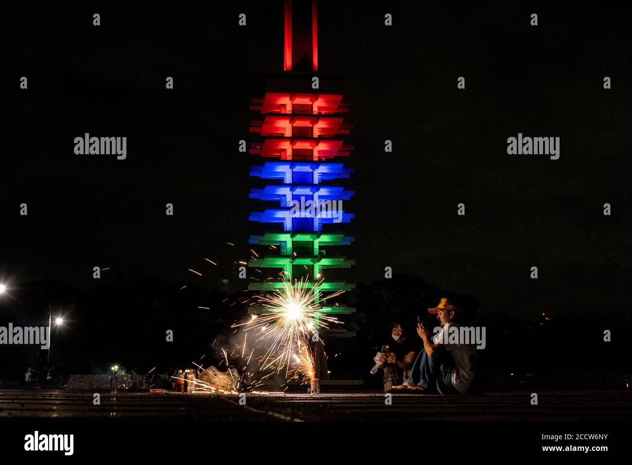 Tokyo, Giappone. 24 Agosto 2020. Una coppia è vista giocare i firecrackers davanti alla Komazawa Park Olympics Memorial Tower è illuminata nel colore delle Paralimpiadi come segna un anno per andare fino a Tokyo 2020 Paralimpiadi. Tokyo 2020 Paralimpiadi e Olimpiadi sono stati rinviati al 2021 a causa della situazione COVID-19. Credit: SOPA Images Limited/Alamy Live News Foto Stock