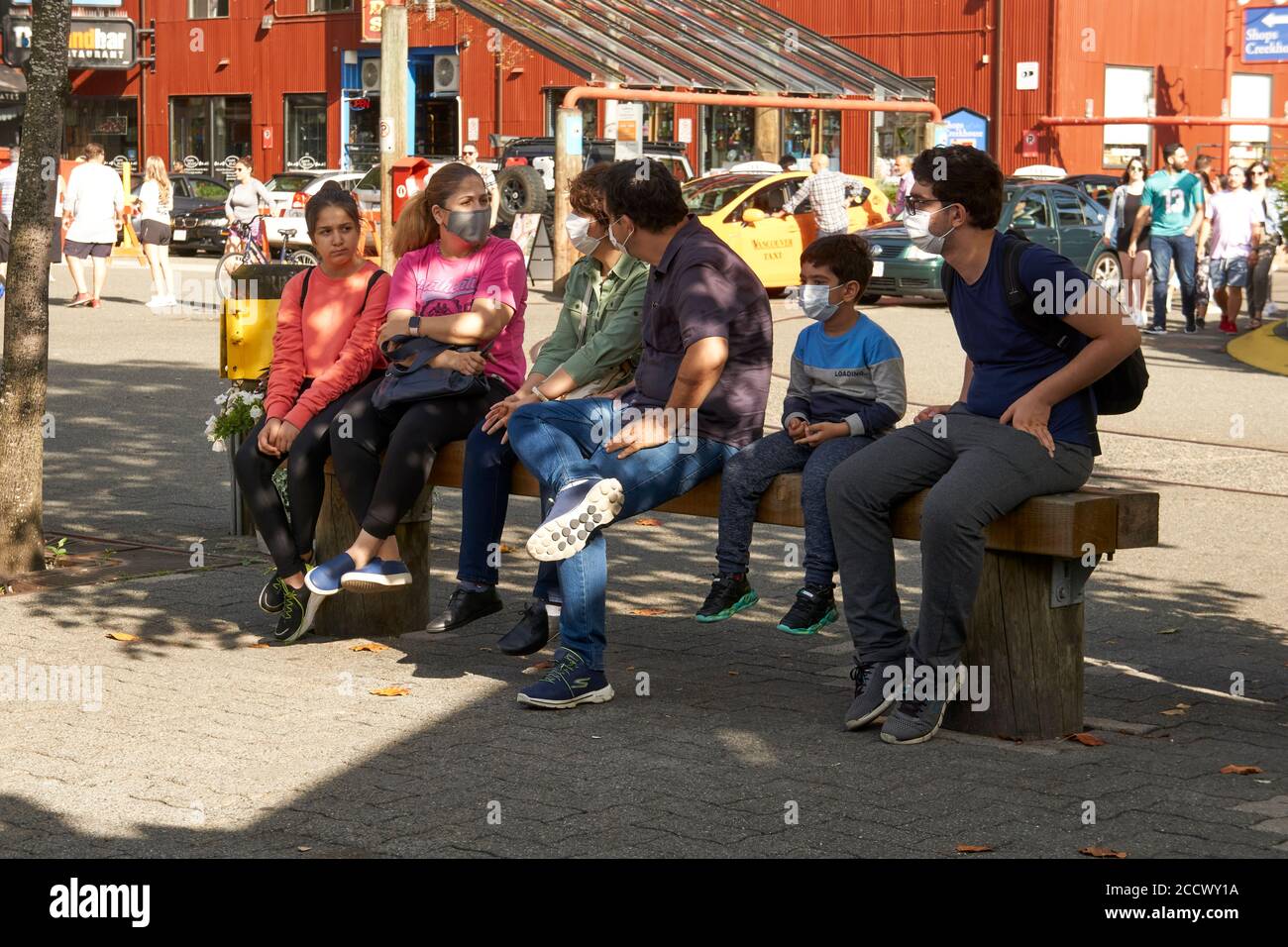 Una famiglia seduta su una panca che indossa maschere facciali protettive all'aperto durante la pandemia COVID19, Granville Island, Vancouver, BC, Canada Foto Stock