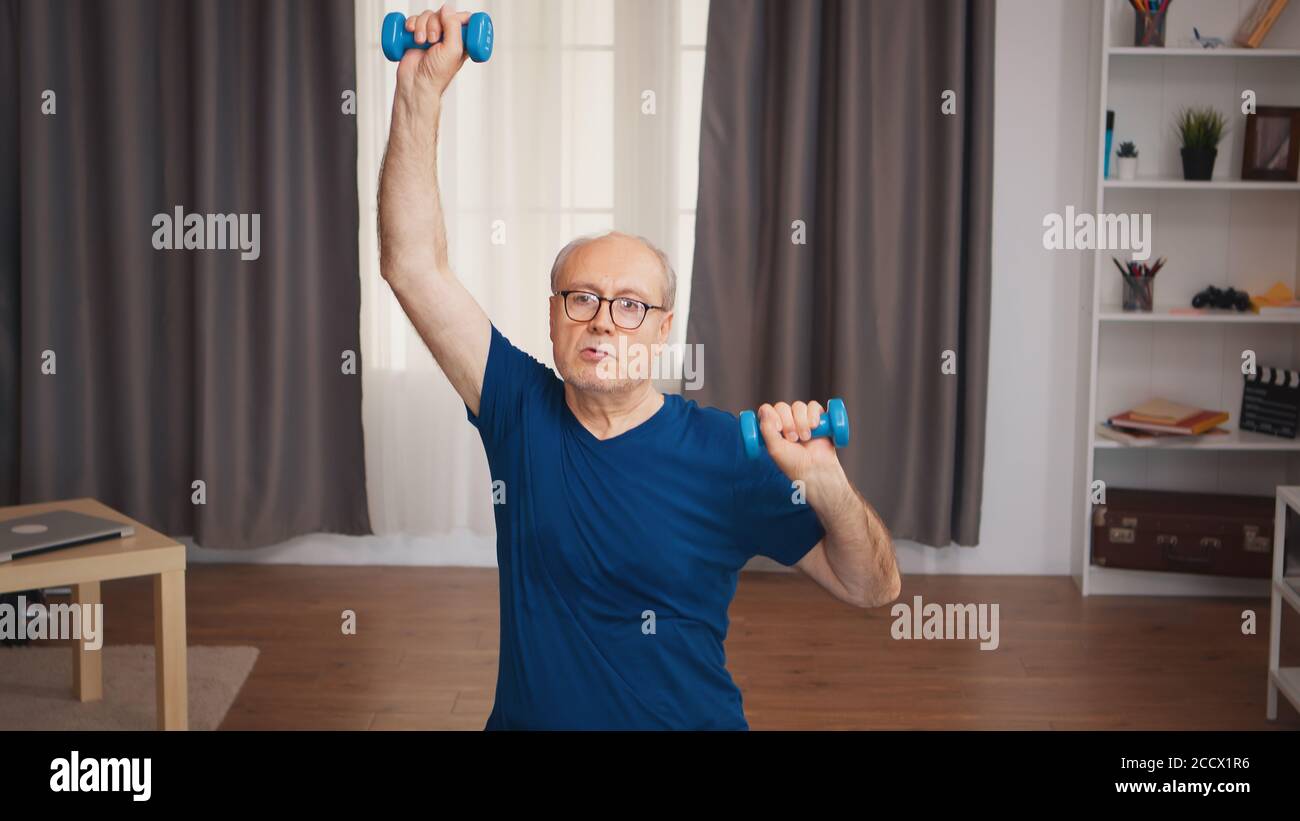 Uomo anziano che fa sport in salotto usando i manubri. Anziano pensionato sano addestrando sport di salute a casa, esercitandosi attività di idoneità all'età anziana Foto Stock