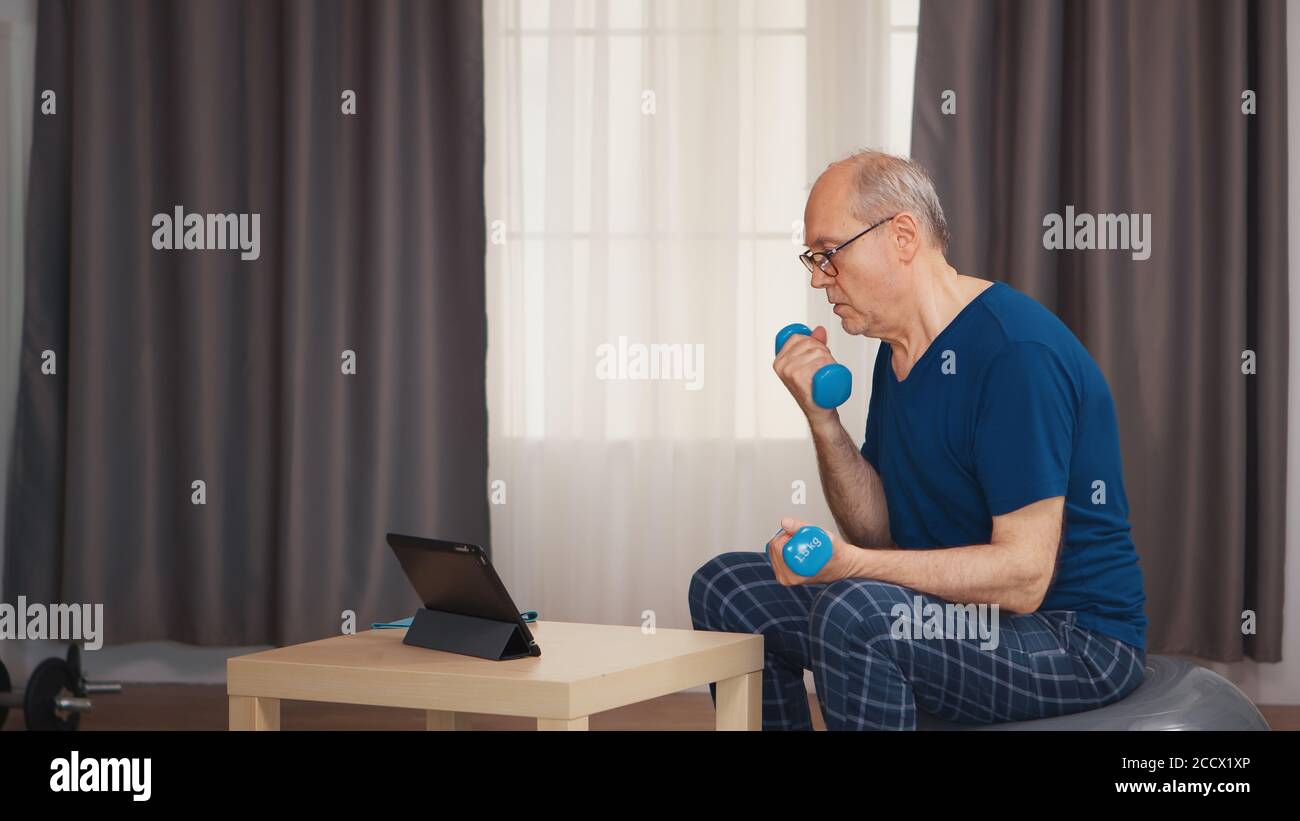 Uomo anziano che si gode di allenarsi con i manubri durante il programma di fitness online. Anziano pensionato sano addestrando sport di salute a casa, esercitandosi attività di idoneità all'età anziana Foto Stock