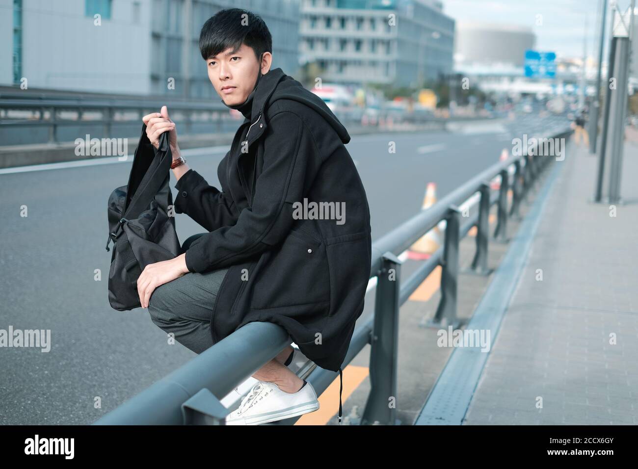 Bello giovane uomo asiatico borsa per il trasporto mentre seduto su recinzione metallica sul sentiero Foto Stock