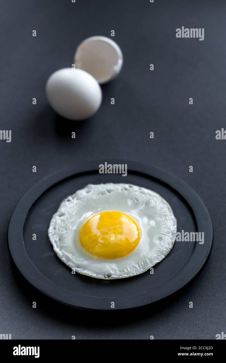 Uova fritte. Primo piano di uova fritte su un piatto nero. Un uovo intero e un guscio accanto ad esso in sfocatura. Su sfondo nero. Messa a fuoco selettiva. Spazio per il testo. Foto Stock