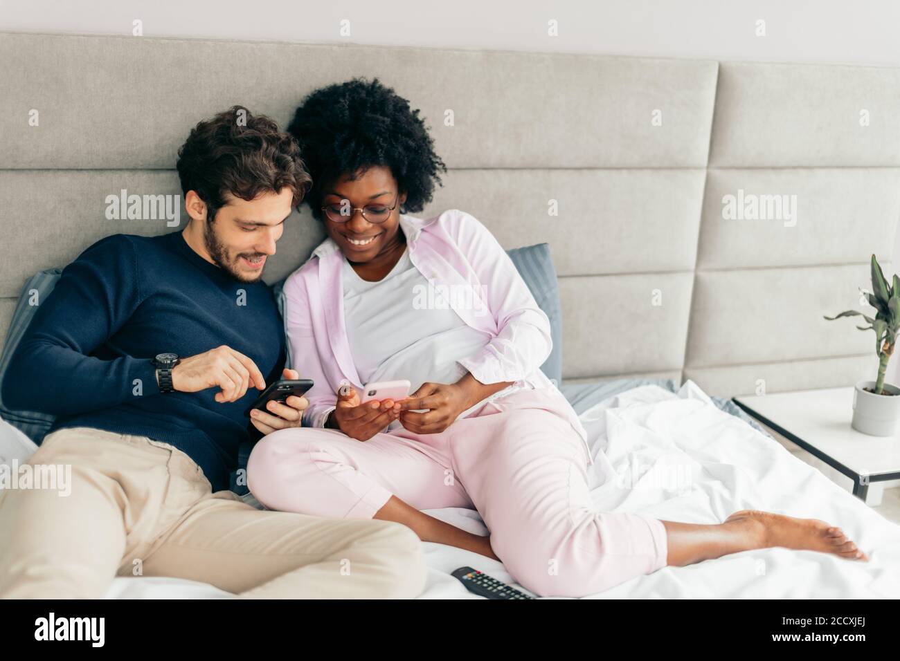 Giovane moglie e marito si rilassano a letto durante la giornata. Spauses di diverse nazionalità che condividono storie divertenti e guardano video con Smart Foto Stock