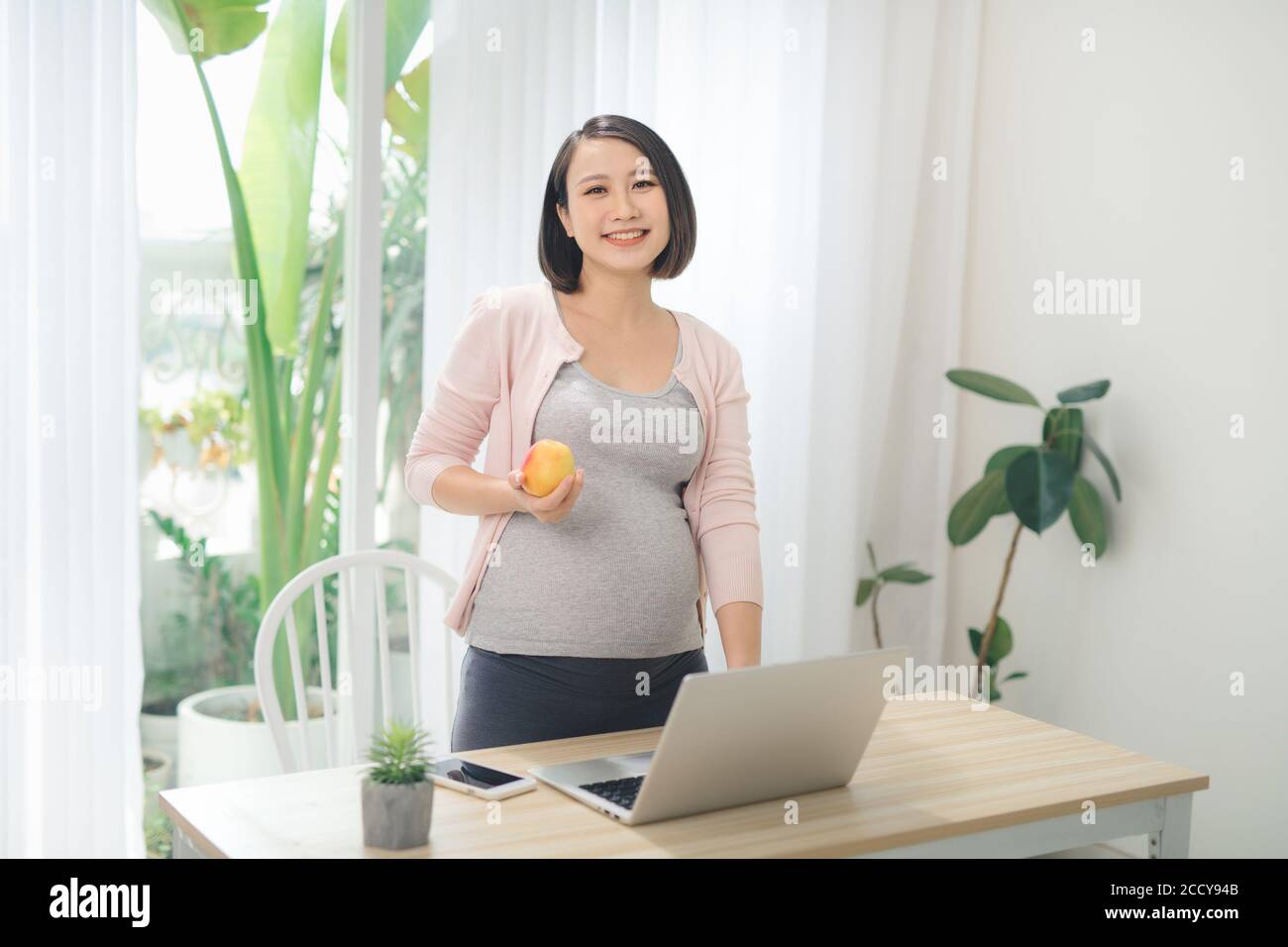 Bella donna d'affari incinta mangiare mela e sorridere alla macchina fotografica Foto Stock