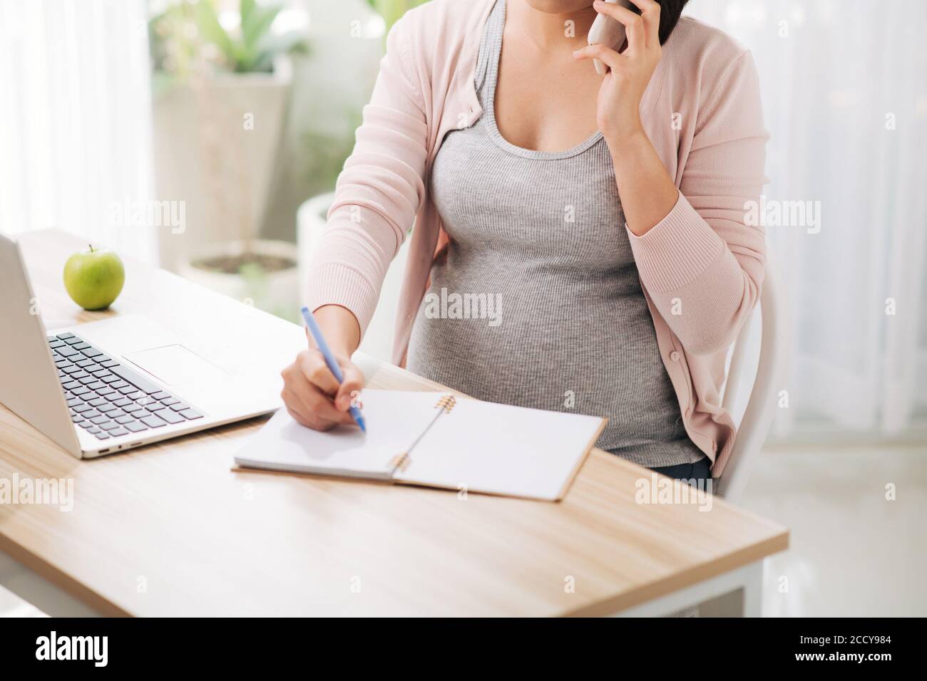 Donna d'affari incinta che parla sul telefono cellulare mentre lavora sul computer portatile in ufficio Foto Stock