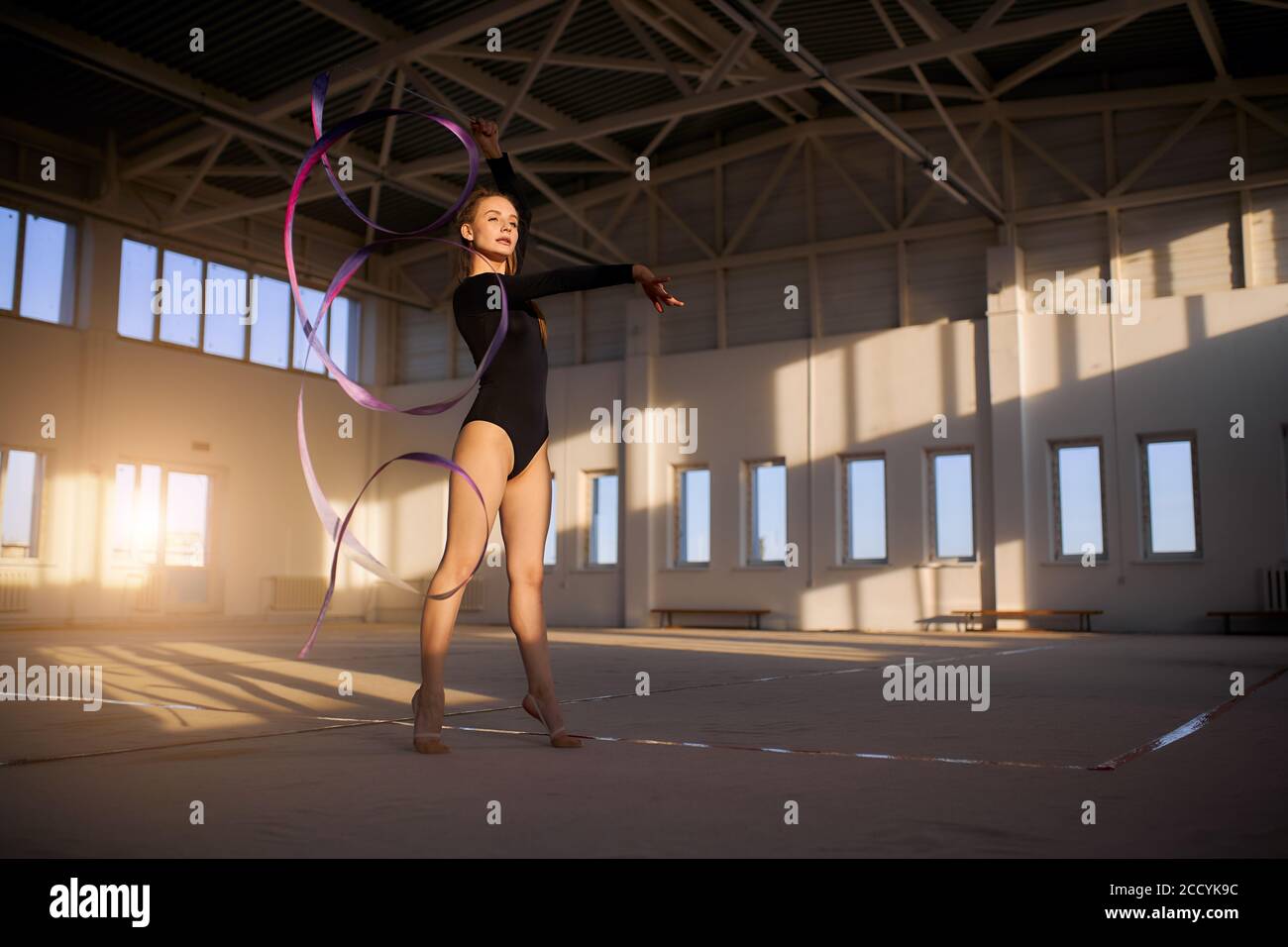 Affascinante bella ragazza allenarsi in nero sport hall, luce giorno che splende la stanza, giocare con nastro lungo, guarda via con espressione fiduciosa, scatto f Foto Stock