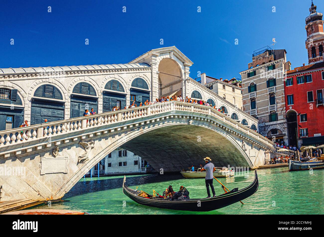 Venezia, Italia, 13 settembre 2019: Gondoliere in gondola tradizionale barca a vela sulle acque del Canal Grande sotto il Ponte di Rialto, tipici edifici colorati e cielo blu chiaro sfondo Foto Stock