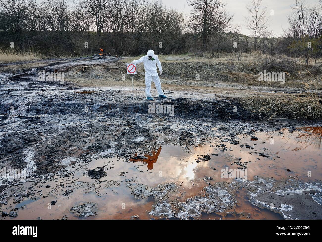 Ricercatore in tuta protettiva bianca che installa un cartello di avvertimento, nessun cartello di ingresso, su territorio contaminato. Acqua fangosa sporca arrugginita per analisi. Concetto di catastrofe ecologica Foto Stock