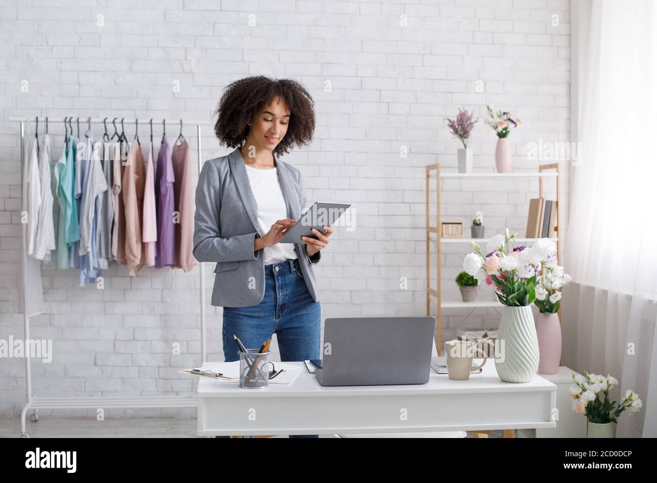 Formazione di design di moda online. Donna afro-americana occupata che tiene il tablet in mano vicino al tavolo con il computer portatile Foto Stock