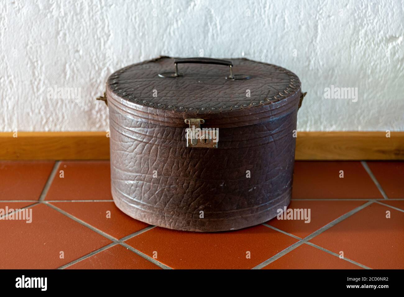 Valigia rotonda vintage adagiata sul pavimento su piastrelle marroni contro una parete bianca. Foto Stock