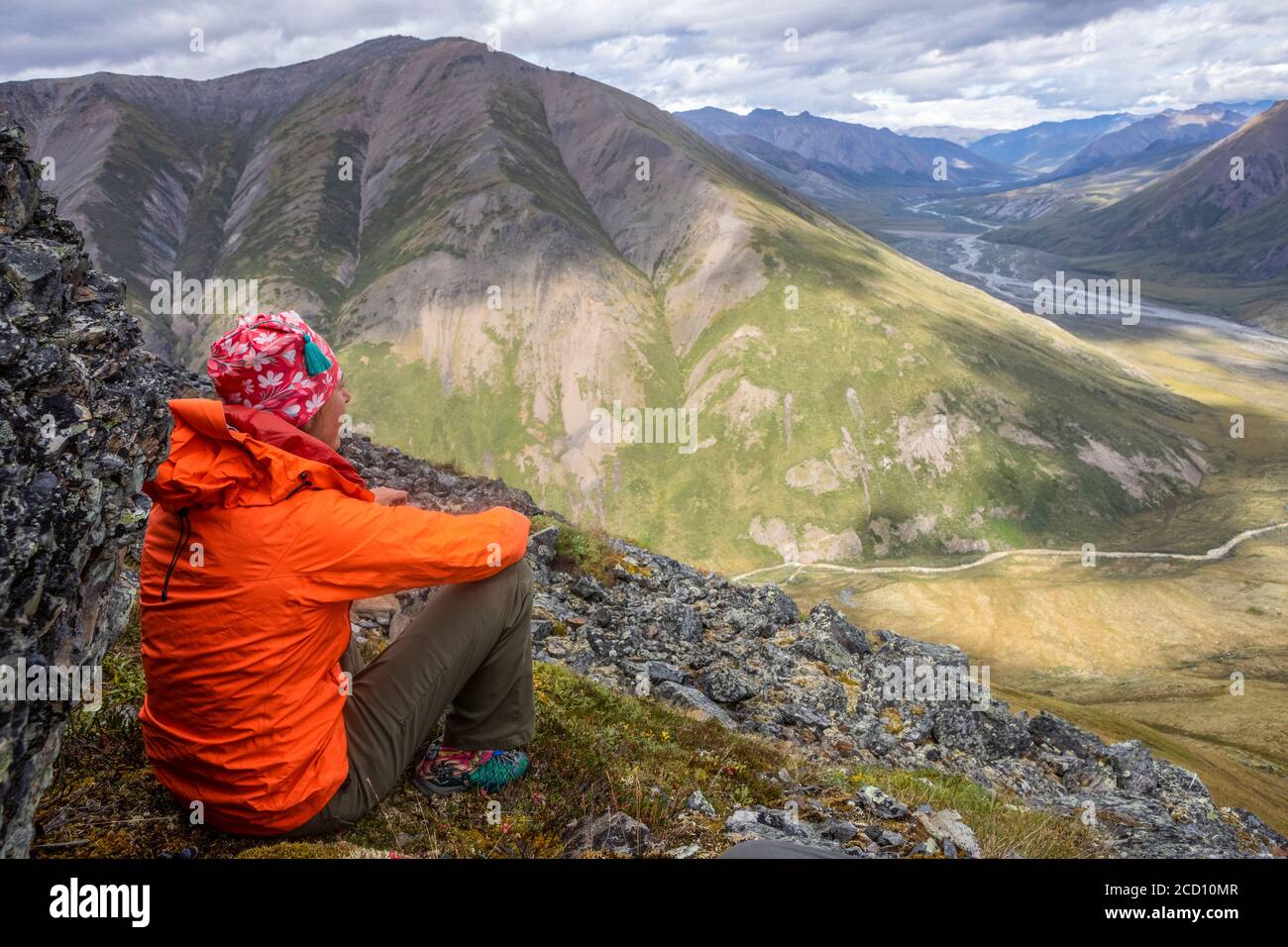 Donna escursionista che indossa giacca da pioggia arancione seduta giù sul fianco della montagna, godendo di vista della valle e insenature e fiumi sotto su una parte di sole, estate ... Foto Stock