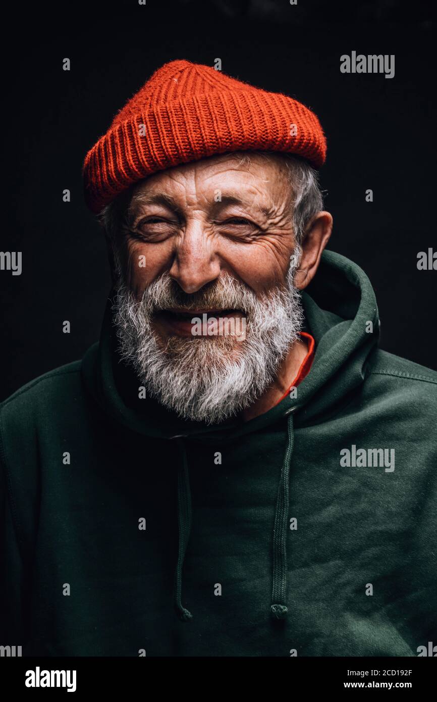 Primo piano ritratto di felice ottimista di 70 anni con volto sorridente e rugoso, vestito con cappello arancione hipster e felpa con cappuccio verde, isolato su nero ba Foto Stock