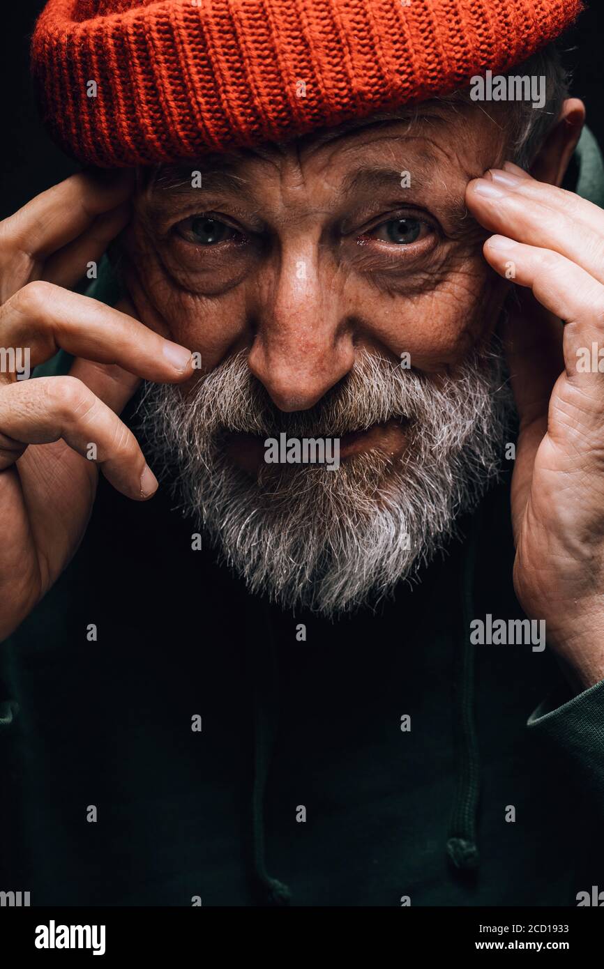 Primo piano ritratto di vecchio uomo bearded in cappello arancione con rughe di viso stagionato stares si spera a fotocamera che tiene le mani vicino ai templi, isolato ov Foto Stock