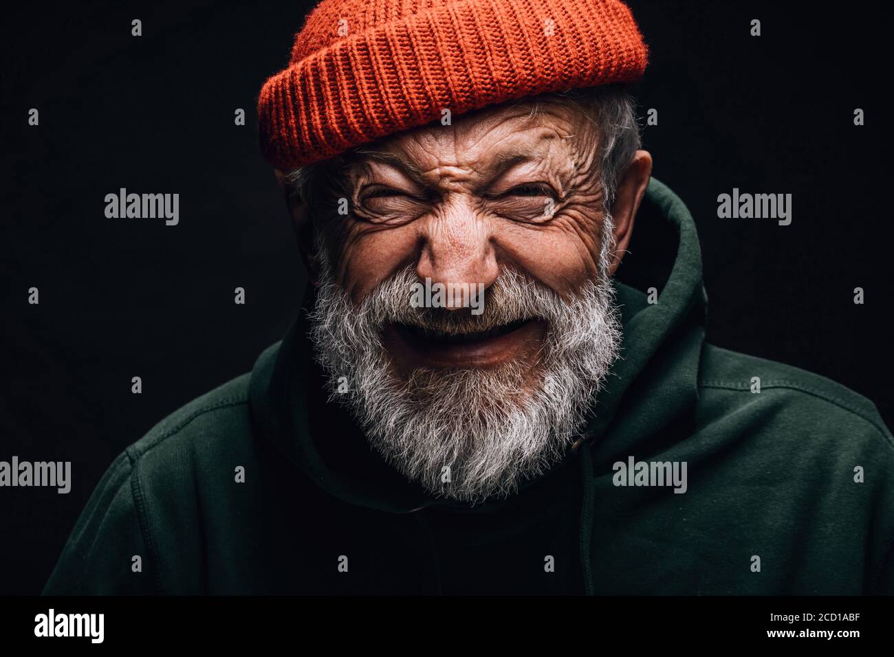 Primo piano ritratto di felice ottimista di 70 anni con volto sorridente e rugoso, vestito con cappello arancione hipster e felpa con cappuccio verde, isolato su nero ba Foto Stock