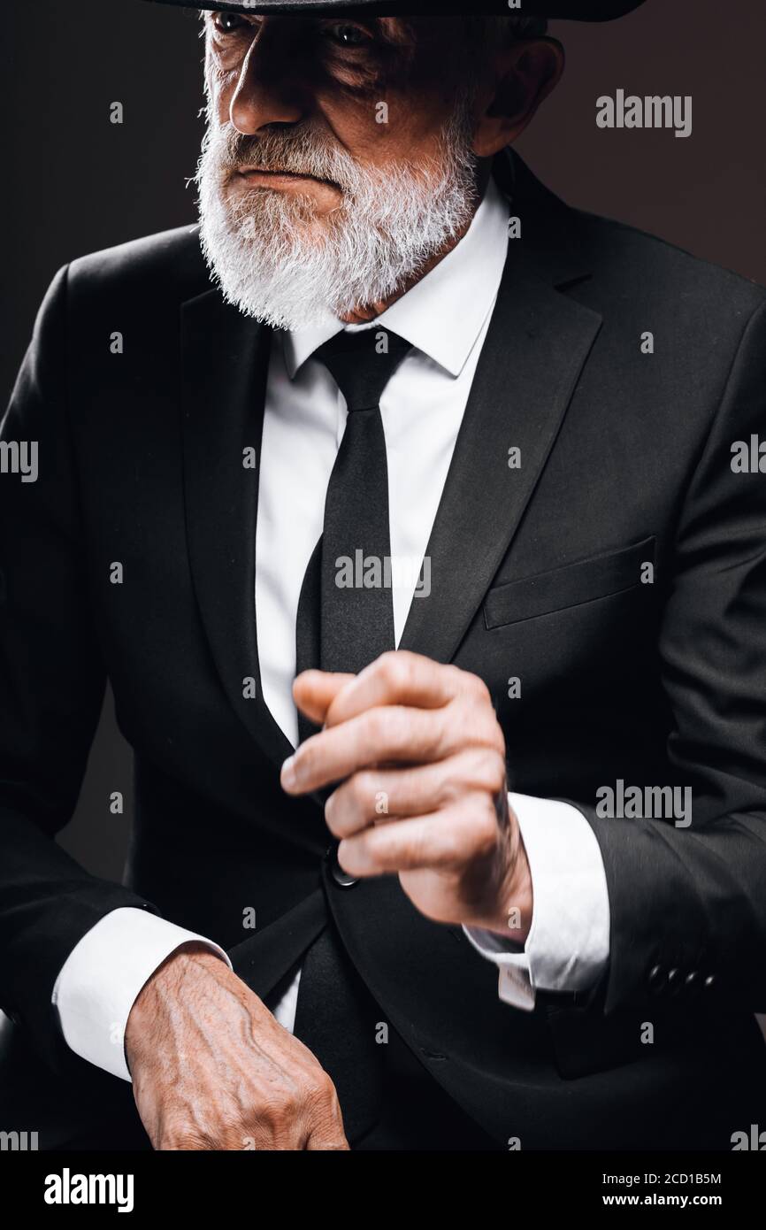 Elegante ritratto per gentiluomo anziano in stile classico con cappello trilby e tuta nera. Moda maschile, look classico concetto. Studio girato al buio Foto Stock