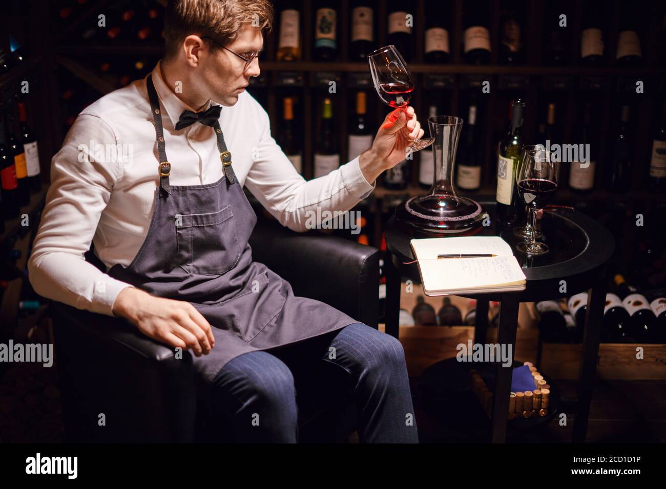 esperienza di degustazione in hotel o ristorante. Ragazzo professionista o sommelier che tiene un bicchiere di delizioso vino rosso che apprecita la sua tinta con bottiglie raccogliere Foto Stock