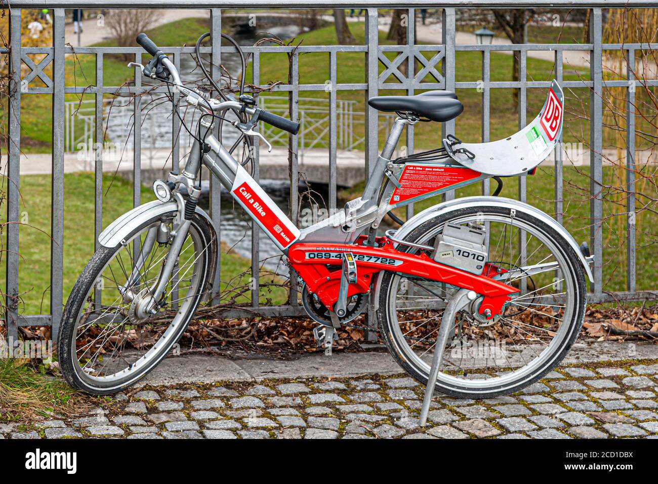 Una bicicletta a noleggio rosso e argento del sistema "Call a Bike" della Deutsche Bahn è bloccata a una recinzione del parco a Monaco di Baviera, Germania, identificabile dal logo DB e dal numero di servizio stampato. Call a Bike è stato lanciato in diverse grandi città nel 2000 ed è uno dei più antichi sistemi di bike-sharing del paese. Foto Stock