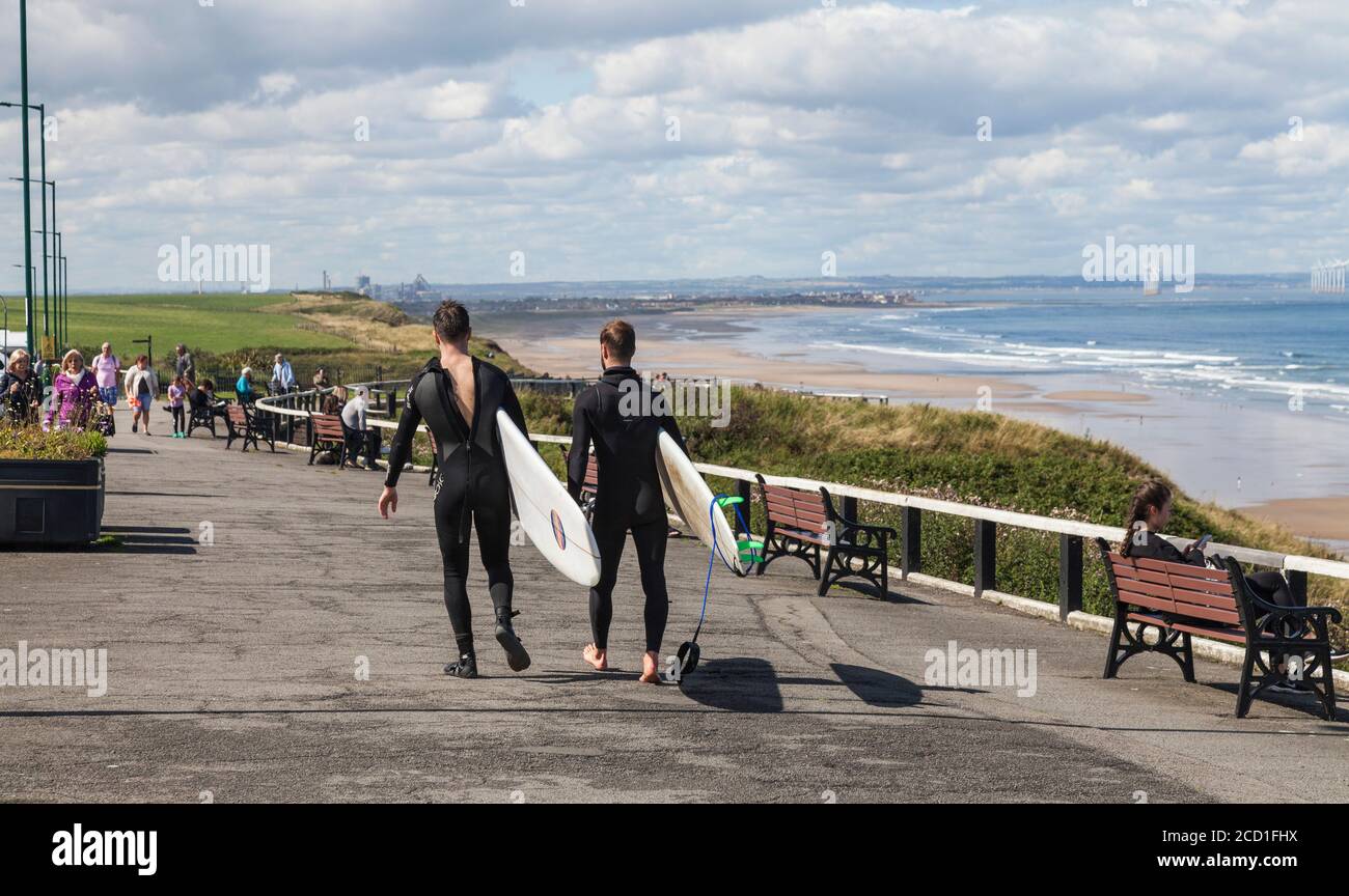 Due giovani uomini in tute umide che trasportano le loro tavole da surf Lungo la passeggiata a Saltburn by the Sea, England, UK Foto Stock